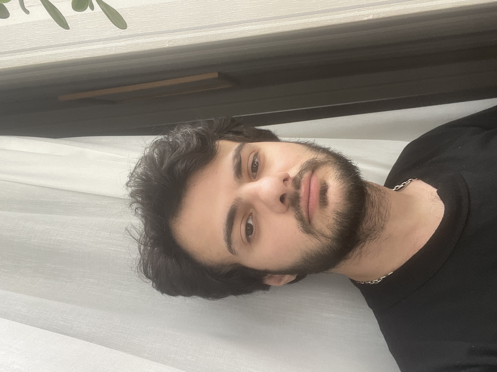 A medium shot, taken from a low angle, shows a young man with dark, curly hair and a beard. He is wearing a black t-shirt and a silver chain. He is looking directly at the camera. The background consists of white sheer curtains and a dark window frame. The lighting suggests natural light, possibly from a window. The image appears to be a selfie.
The photo was taken indoors, likely in a residential setting, in Tbilisi, Georgia, on an overcast day, evidenced by the diffused natural light coming from a window. The time of day is likely daytime due to the presence of natural light.