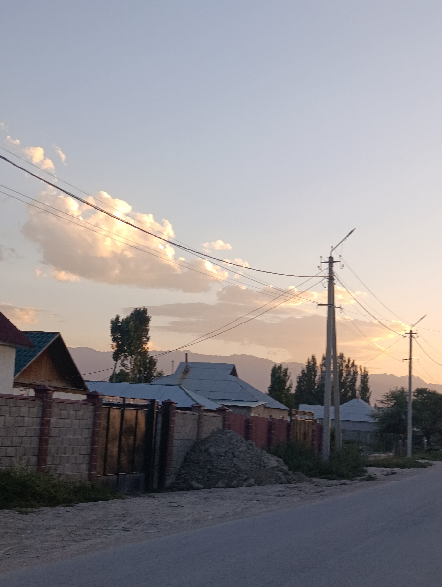 The image depicts a street scene in the city of Kochkor, Kyrgyzstan, at dusk. The sky is a soft gradient of pale blue and warm orange, with scattered clouds illuminated by the setting sun. Power lines stretch across the frame, supported by utility poles. In the foreground, a paved road runs parallel to a brick wall with a metal gate. Beyond the wall, several houses with pitched roofs are visible, interspersed with trees and mountains in the distance. A pile of construction debris sits to the right of the gate. The overall atmosphere is peaceful and tranquil, characteristic of a quiet evening in a rural or semi-rural setting.