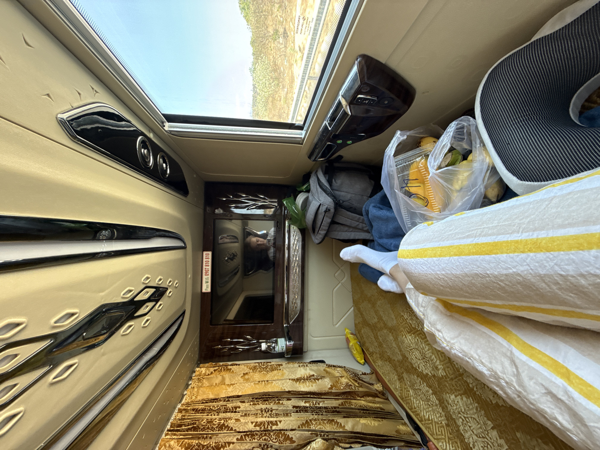 This image provides an interior view of a luxurious and modern sleeper bus compartment, likely in Phuoc Huu, Vietnam, designed for comfortable long-distance travel. The scene is illuminated by natural daylight streaming through a large window or sunroof overhead, revealing a clear blue sky and a glimpse of the passing landscape with distant trees.
The central subject is a person resting or reclining within the private berth. Their lower body, clad in white socks, is partially covered by a white and yellow striped blanket or sheet. A patterned gold mattress or cushion forms the base of the sleeping area. A grey and black patterned neck pillow is visible near the head.
The compartment features a sophisticated design with beige and tan upholstered walls, complemented by dark wooden accents and shiny metallic trim. On the left wall, a control panel with two circular dials and a decorative metallic inlay is visible. Further along this wall, a dark wooden unit with a reflective surface reflects the upper body and face of the resting person. A sticker on this unit displays text: "Pass Wifi: 0907 810 810". A small water bottle is held in a holder beneath this unit.
Near the head of the berth, various personal items are stowed: a clear plastic bag filled with fruits (bananas and possibly others), a grey backpack, and a blue fabric item. Another control panel with multiple buttons and indicators, featuring a dark wooden finish, is located on the right side of the compartment, above the head area.
The overall impression is one of relaxed and convenient travel, characteristic of modern sleeper buses in Southeast Asia.