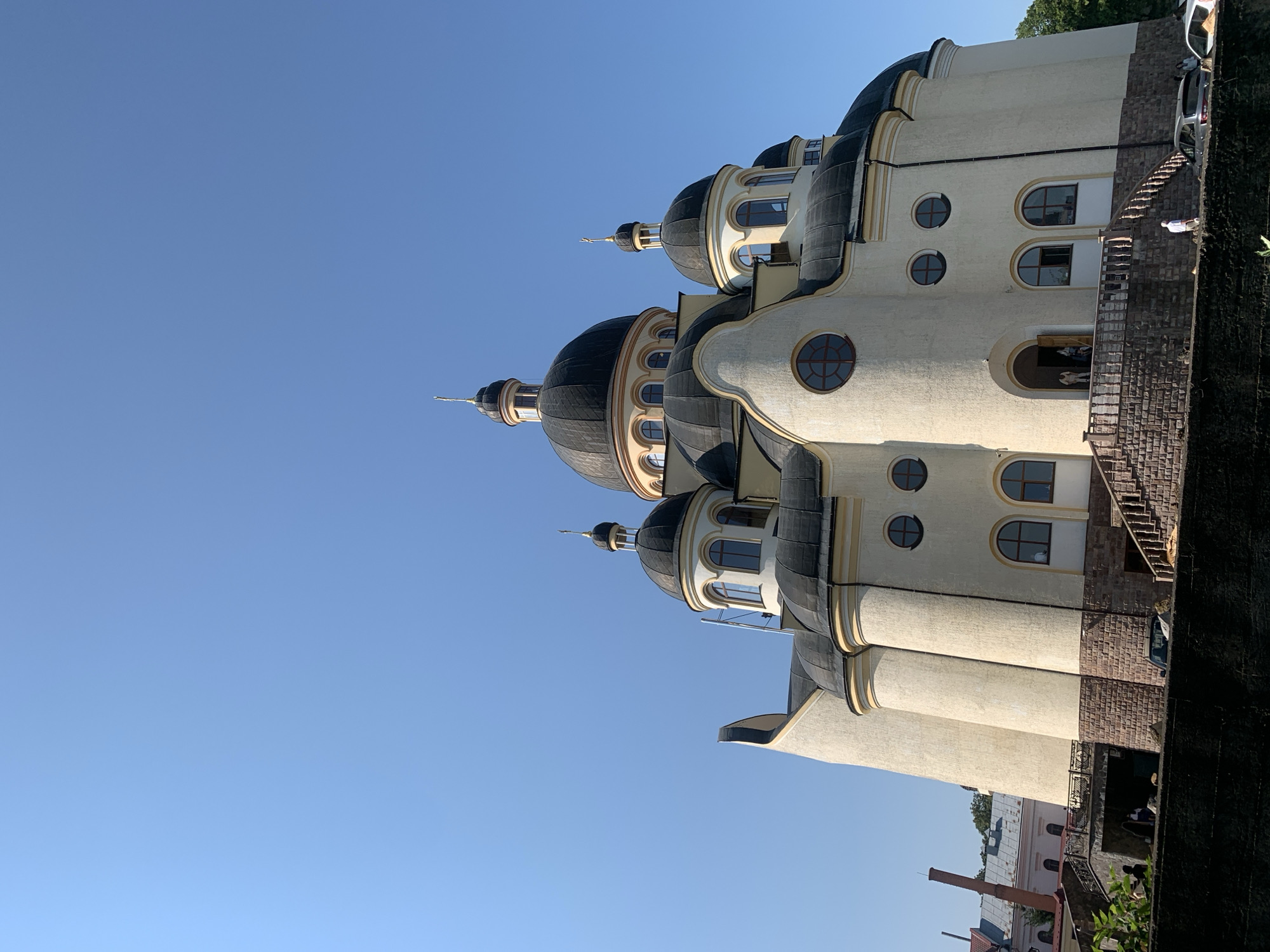 A large, cream-colored church with a black roof and multiple domes stands against a clear blue sky. The church has a grand entrance with stone steps leading up to a doorway where several people are visible. The architecture is ornate, with arched windows and decorative details. In the foreground, a dark wall runs across the bottom of the image, and a few cars are parked near the entrance. To the left, a tall brick chimney rises behind a building. The overall impression is of a bright, sunny day in a European city, likely Kolomyia, Ukraine, given the architectural style.