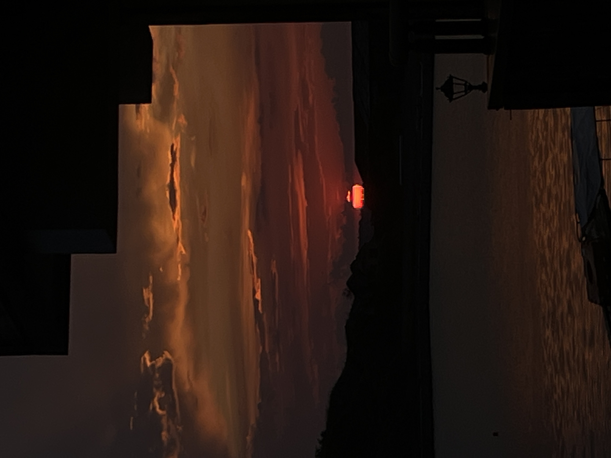 The image captures a dramatic sunset or sunrise over a body of water, framed by dark architectural elements. The sky is ablaze with intense orange, red, and golden hues, with clouds reflecting the vibrant light. A distinct, glowing red orb, representing the sun, is visible just above a dark, silhouetted mountainous or landmass horizon.
In the foreground, on the left, a dark, angular structure, possibly part of a building or a balcony, partially obstructs the view of the sky. On the right, another dark, silhouetted structure, likely a building, features a traditional-style black metal lantern hanging from its side. Below these foreground elements, a body of water, appearing as a lake or sea, reflects the warm colors of the sky with subtle ripples. The scene suggests either dusk or dawn, given the low position of the sun and the rich, warm light bathing the clouds. There are no people visible, nor any discernible text.
