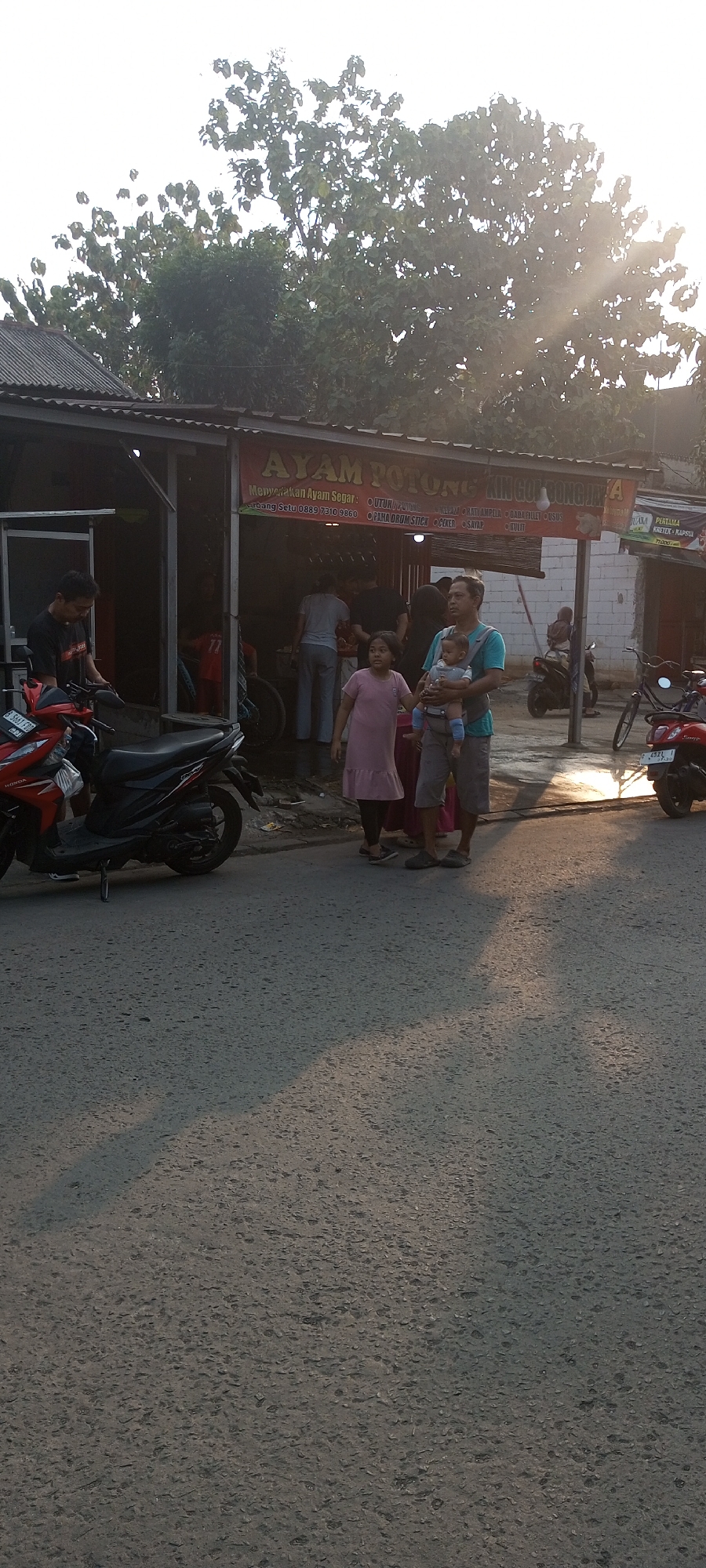The image depicts a street scene in Setu, Indonesia, in front of a food stall named "AYAM POTONG KING GULI BONGHATA" which advertises fresh cut chicken. The stall has a corrugated metal roof and a red awning. Several people are present. A man in a black t-shirt stands by a red motorcycle parked on the left side of the road. In front of the stall, a family is gathered. A man in a blue shirt and grey shorts is holding a baby in a carrier, and standing next to him is a young girl in a pink dress. Behind them, several other people are visible inside and around the food stall. The road is paved and has distinct shadows cast by trees and buildings. A bicycle and another motorcycle are also visible in the background. The lighting suggests it is late afternoon or early evening, with the sun casting long shadows.