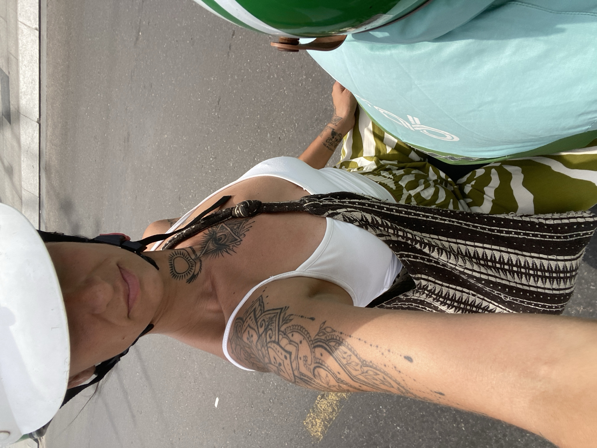 A woman wearing a white helmet and a white tank top rides a scooter on a paved road in Phu Thuy Ward, Vietnam. She has tattoos on her neck and arm, and a patterned bag hangs from her shoulder. The road has yellow markings. A person in a green and white helmet is visible to the right of the frame.