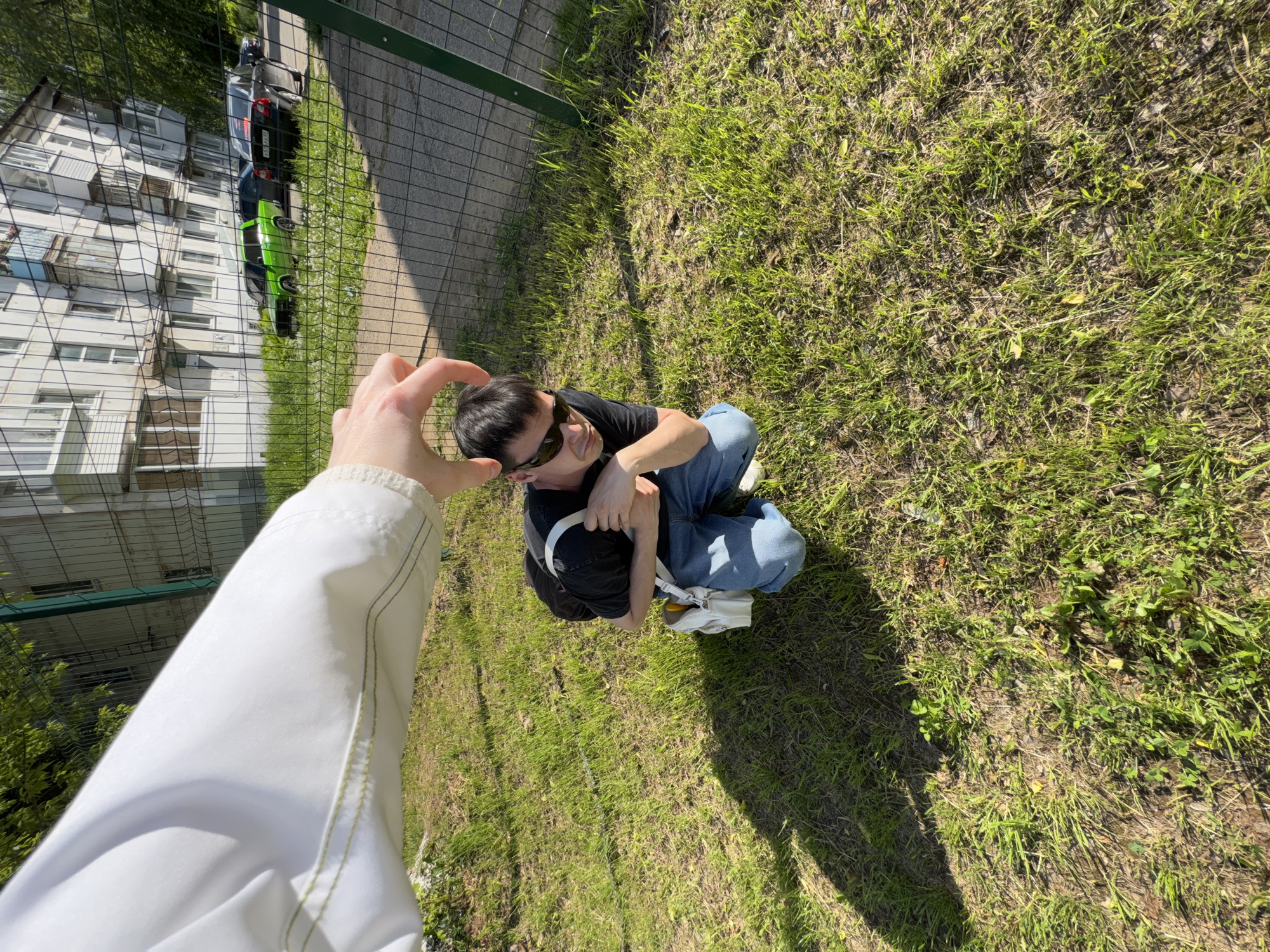 A person kneels on grassy ground, looking up and to the left. A second person's arm and hand reach down from the upper left of the frame, positioned as if framing the kneeling person's face. The kneeling person wears sunglasses, a dark shirt, blue jeans, and white sneakers. The grass appears to be located next to a sidewalk and a chain-link fence, behind which is a multi-story building and a couple of vehicles. This photograph was taken in the city of Chaikovsky, Russia.