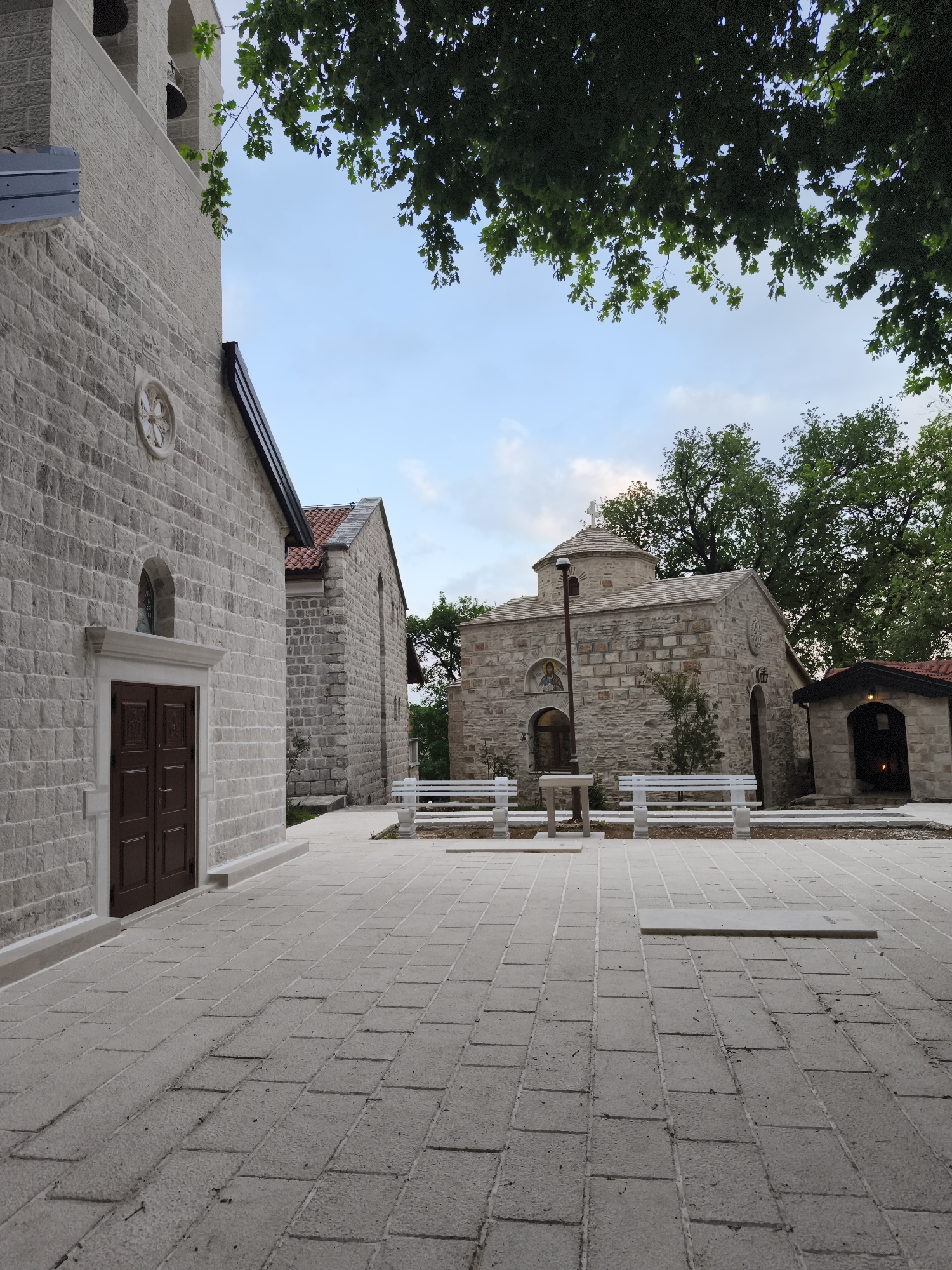 The image captures a courtyard setting, dominated by grey stone buildings typical of the architecture in the city of Budva, Montenegro. The stonework exhibits a regular, block-like pattern with minimal ornamentation.
A church building with a red-tiled roof is partially visible in the background. A smaller chapel-like structure with a single dome is positioned centrally, featuring a small religious icon above its entrance. There are white benches flanking a water fountain or baptismal font in front of the chapel.
The foreground is paved with stone bricks and leads to the main church building with a prominent, dark brown wooden door. A bell tower is attached to the church. Overhanging tree branches are visible at the top of the frame. The sky is a soft blue with scattered clouds. The scene is bathed in even daylight, suggesting it might be a late afternoon or early morning shot. The overall impression is of a tranquil and well-maintained religious site. No people are visible.