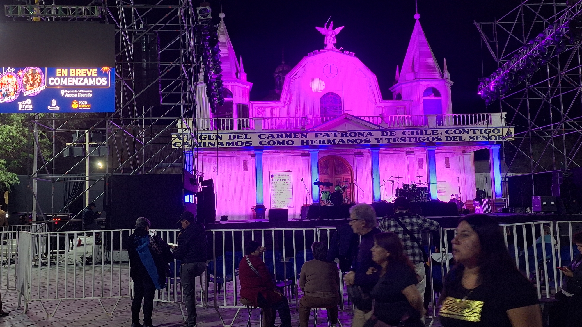 The image depicts an outdoor evening event in Pozo Almonte, Chile. A large stage with musical equipment, including drums and microphones, is set up in front of a building illuminated by vibrant purple and pink lights. The building appears to be a church, with architectural features like spires and columns visible. A banner across the front of the building reads "VIRGEN DEL CARMEN PATRONA DE CHILE, CONTIGO CAMINAMOS COMO HERMANOS Y TESTIGOS DEL SEÑOR." To the left of the stage, a large LED screen displays "EN BREVE COMENZAMOS" (We begin shortly) along with logos, suggesting an organized public gathering.
A metal scaffolding structure, likely for lighting and sound equipment, is prominently positioned on the left side of the frame, extending upwards and partially obscuring the screen and the sky. In the foreground, a white metal fence separates the audience area from the stage. Several individuals are visible, some standing and observing the scene, while others are seated behind the fence. Their attire suggests casual evening wear. The overall atmosphere appears festive and anticipatory, with the lighting creating a dramatic and engaging visual spectacle. The presence of the screen and stage setup indicates a planned event or performance is about to commence.