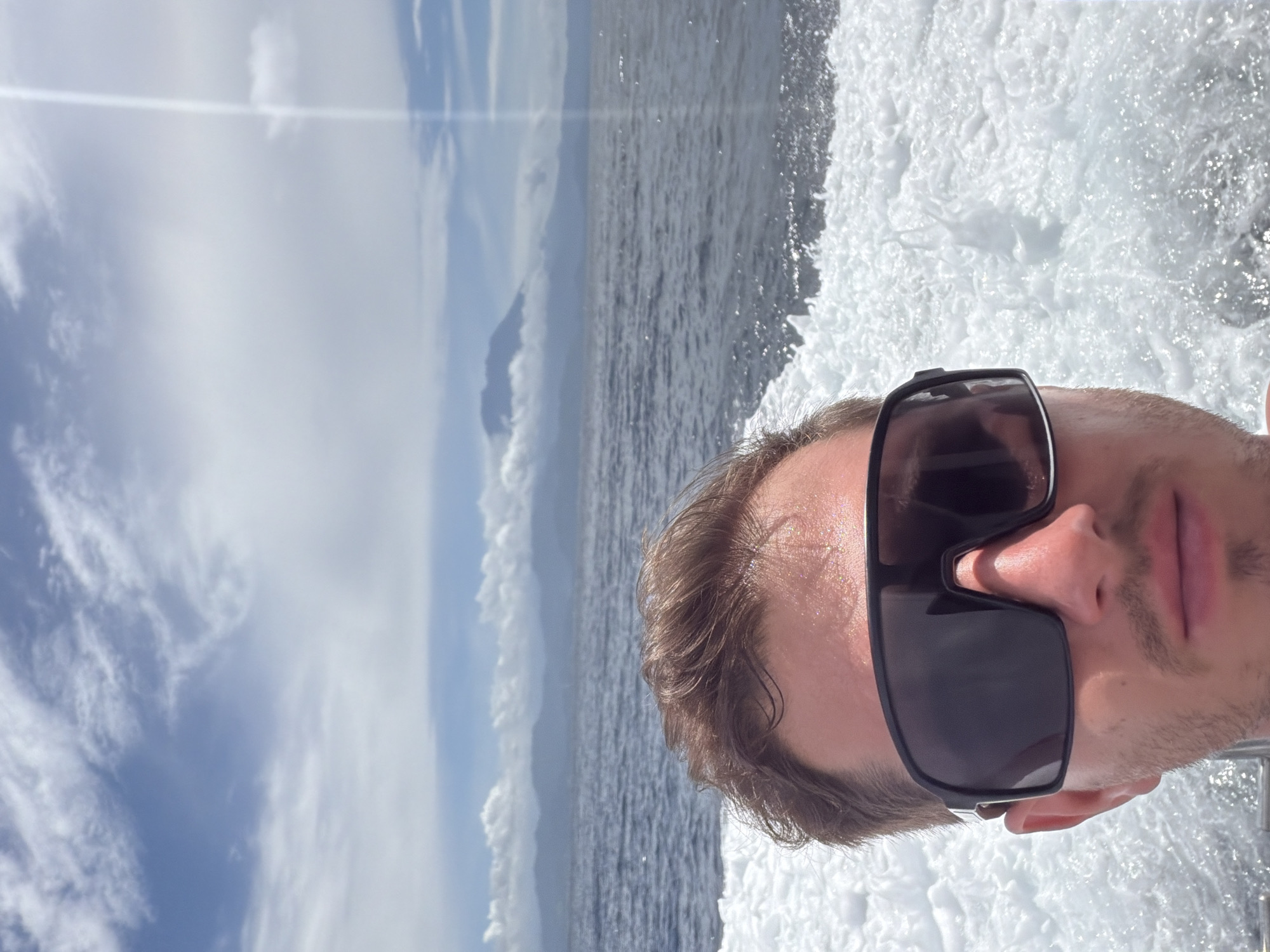 This image, when correctly oriented, is a close-up selfie-style shot of a person on a boat, captured against a scenic ocean and sky backdrop. The primary subject is a male, whose face is visible from the upper lip to the hairline. He is wearing large, dark, rectangular sunglasses that completely obscure his eyes. His hair is brown, and he has light stubble on his chin and jawline. The expression on his visible face appears neutral.
To the right of the subject, a significant portion of the frame is filled with frothy, white, churning water, indicative of a boat's wake. This suggests the person is on a fast-moving vessel. The white foam contrasts with the deeper blue of the surrounding sea.
In the background, to the left of the subject, the expansive ocean stretches towards the horizon under a bright blue sky. A distinct landmass, likely a mountain or island, is visible in the distance, partially shrouded by clouds. The sky itself features scattered white clouds, and a faint horizontal light streak, possibly a lens flare, crosses the upper portion of the image.
The scene conveys a bright, sunny daytime setting, suggesting favorable weather conditions for being out on the water. The activity is boating or a similar water excursion. The location is Nusa Penida, Indonesia. No visible text is present in the image.