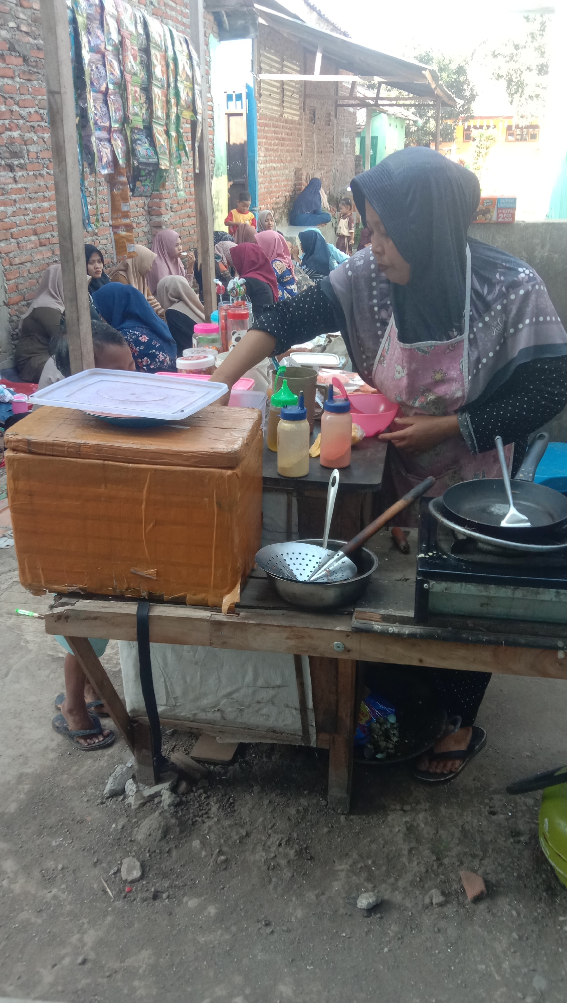 In Karang Anyar, Indonesia, a food stall is set up outdoors, likely on a street or in a market. A woman wearing a dark hijab and a patterned apron is the central figure, preparing food. She is standing behind a wooden table laden with cooking equipment and ingredients. To her right, a small black gas stove has a pan on it, and a metal spatula rests on the edge of the stove. In front of the stove is a metal basin containing a perforated metal strainer with a wooden handle. To the left of the stove are several bottles, including two squeeze bottles with red and yellow caps, and a taller yellow bottle. A pink bowl is also visible on the table. To the left of these items, a large, light brown cardboard box, possibly a cooler, is covered by a white plastic lid. Behind the table, several people are seated, some wearing hijabs. A brick wall forms the backdrop, with packets of goods hanging from a wooden beam above. The ground is dirt and gravel. The scene appears to be a casual, open-air food preparation and service area.
