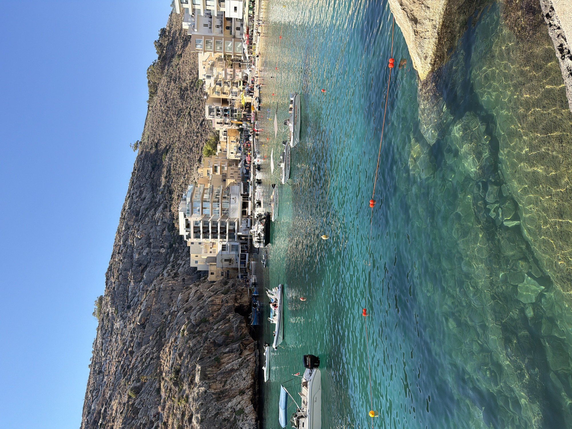 This is a landscape photograph of a picturesque bay in Xlendi, Malta, taken on a clear, sunny day. The image is oriented vertically, with the sky on the left and the sea on the right.
The scene features a rocky cliff face on the left, partially covered in dry vegetation, with a cluster of white and beige buildings built into the hillside on the right. The buildings have multiple stories and balconies, indicative of a coastal town.
At the base of the cliff and extending into the water is a beach area with several people sunbathing and walking. The clear, turquoise water of the bay is a prominent feature, with several boats of various sizes anchored or moored. Some of these boats appear to be small motorboats and a larger inflatable boat, with people visible on board some of them. A rope with orange buoys stretches across a section of the water, likely marking a swimming area. The water is shallow enough in some areas to see the rocky seabed and submerged rocks.
The bright sunlight suggests it is midday. The overall atmosphere is one of a tranquil and sunny summer day at a popular seaside resort.