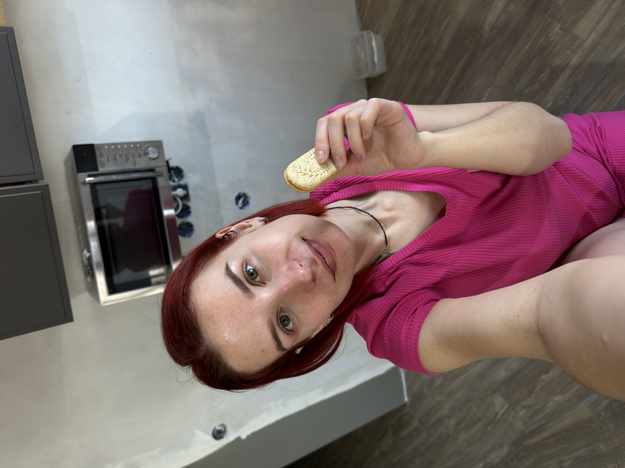 A red-haired woman in a pink ribbed shirt holds a rectangular cookie in her left hand in a kitchen. An oven with a dark interior is visible against a light grey wall with unpatched holes. A grey cabinet is to the left of the oven. Above and behind the woman is a dark wood-patterned floor. A clear plastic container is also visible above and behind the woman. The photo was likely taken in the city of Chaikovsky, Russia.