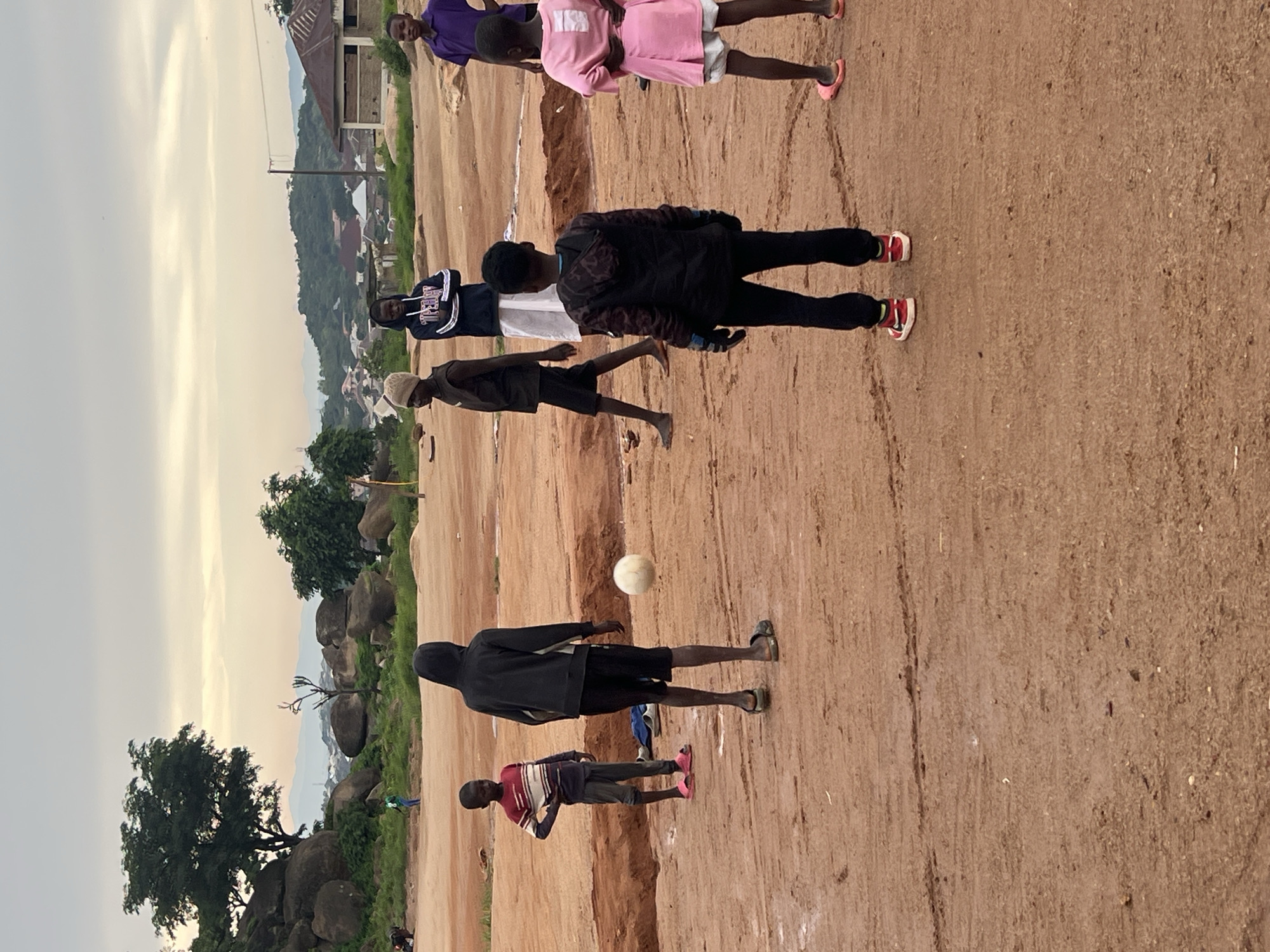 A group of seven children are playing soccer on a dirt field in Jos, Nigeria. The sky is overcast and the ground is a reddish-brown dirt. In the background, there are rocky hills, trees, and buildings. The children are of various ages and are wearing casual clothing. One child is wearing a pink dress, another a purple shirt, and the others are in t-shirts and shorts. One child is wearing a hooded jacket. A soccer ball is in the air, suggesting the game is in progress. The overall scene depicts an outdoor recreational activity in a semi-urban environment.