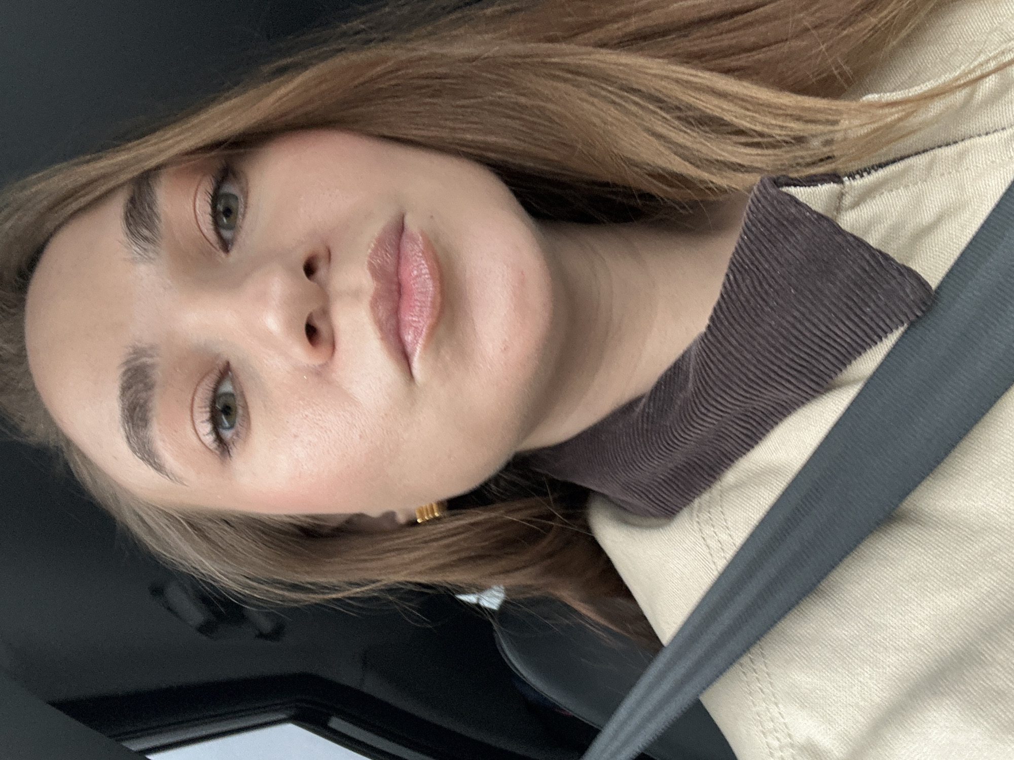 A close-up selfie of a fair-skinned woman with light brown hair and green eyes, wearing a light-colored jacket with a brown collar and a gray seatbelt. The background suggests she is in a car. The location is not visible and therefore unknown.