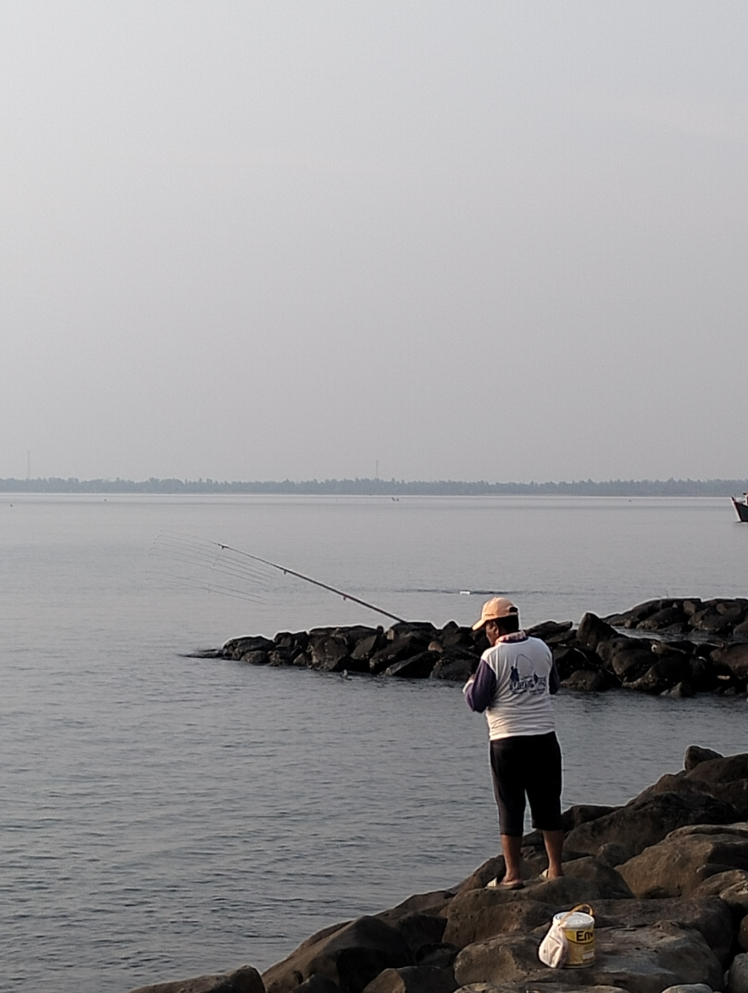 A man is fishing on a rocky shoreline in Lhokseumawe, Indonesia. He wears a light-colored hat, a white and purple long-sleeved shirt, and dark shorts. The man stands on the rocks facing the water, holding a fishing rod. Several fishing rods are visible extending from the rocks into the water. A bucket and a bag are located on the rocks near the man. In the distance, a flat coastline is visible under a grey sky. The sea is calm.