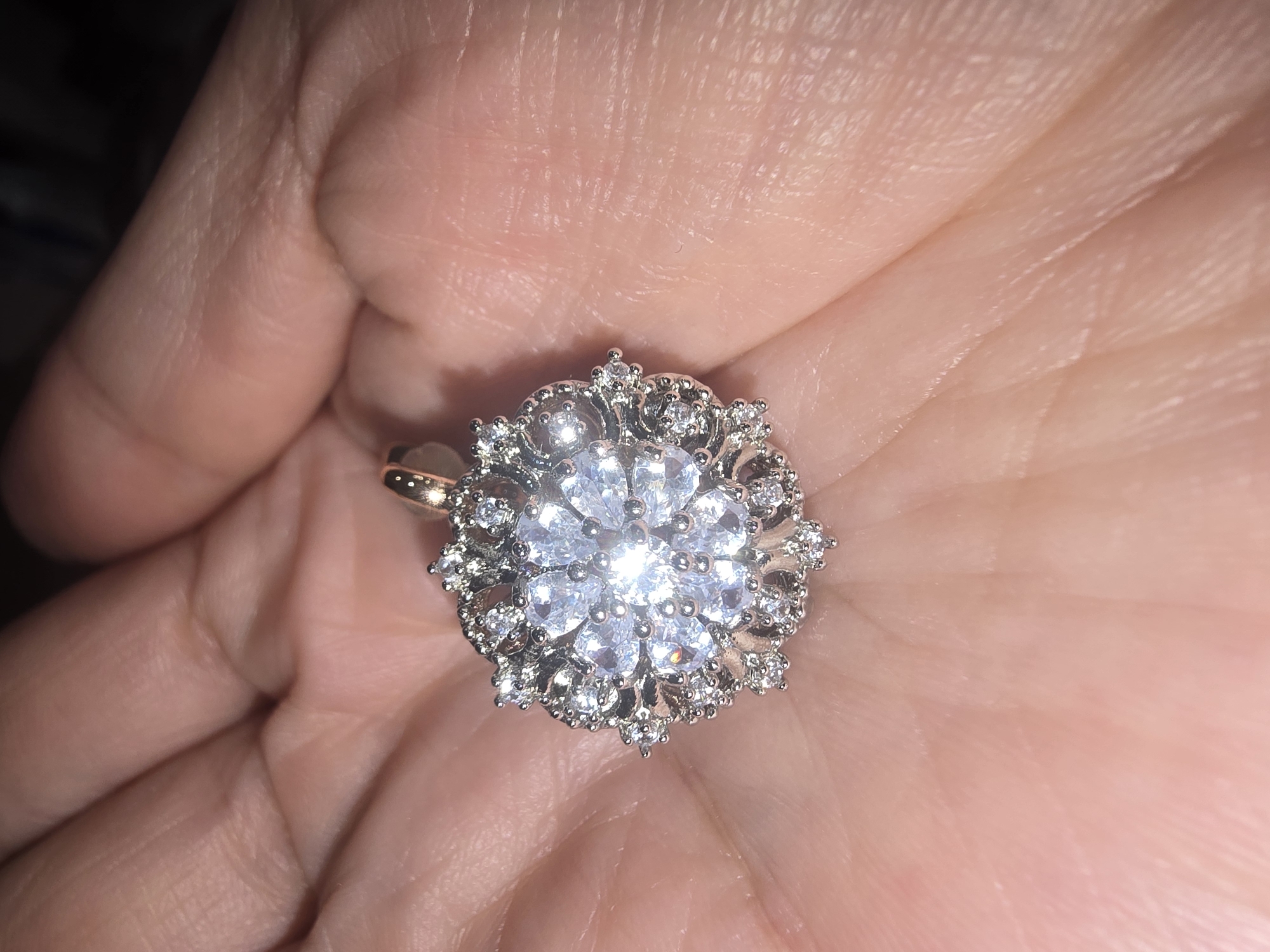 A hand holds a circular, snowflake-shaped pendant adorned with numerous clear stones. The pendant has a silver-toned metal setting and a small rose-gold loop at the top for attachment. The stones are arranged in a multi-layered floral or starburst pattern, with larger stones at the center and smaller ones radiating outwards. The hand holding the pendant is light-skinned and shows visible lines and texture. The background is dark and out of focus. There is no discernible information about the location in Ulan-Ude, Russia.