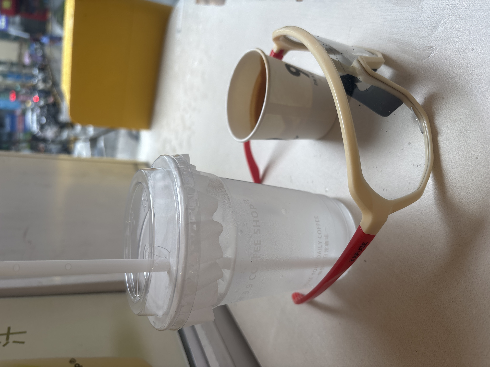 A close-up, eye-level, indoor shot shows a clear plastic cup with a white straw filled with a clear liquid, resting on a white surface. The cup has water droplets on its exterior and branding that includes "9 COFFEE SHOP" and what appears to be Chinese characters. To the right of the cup is a small, white paper cup containing a brown liquid, also with some branding, including the numeral "9". In the foreground, a pair of large, beige-framed sunglasses with red accents on the arms is placed horizontally, partially obscuring the base of the plastic cup. The background is blurred, suggesting an urban setting with buildings, vehicles, and traffic lights. The overall scene depicts a casual moment with beverages and eyewear, likely in a cafe or similar establishment in Nanhai District, China.