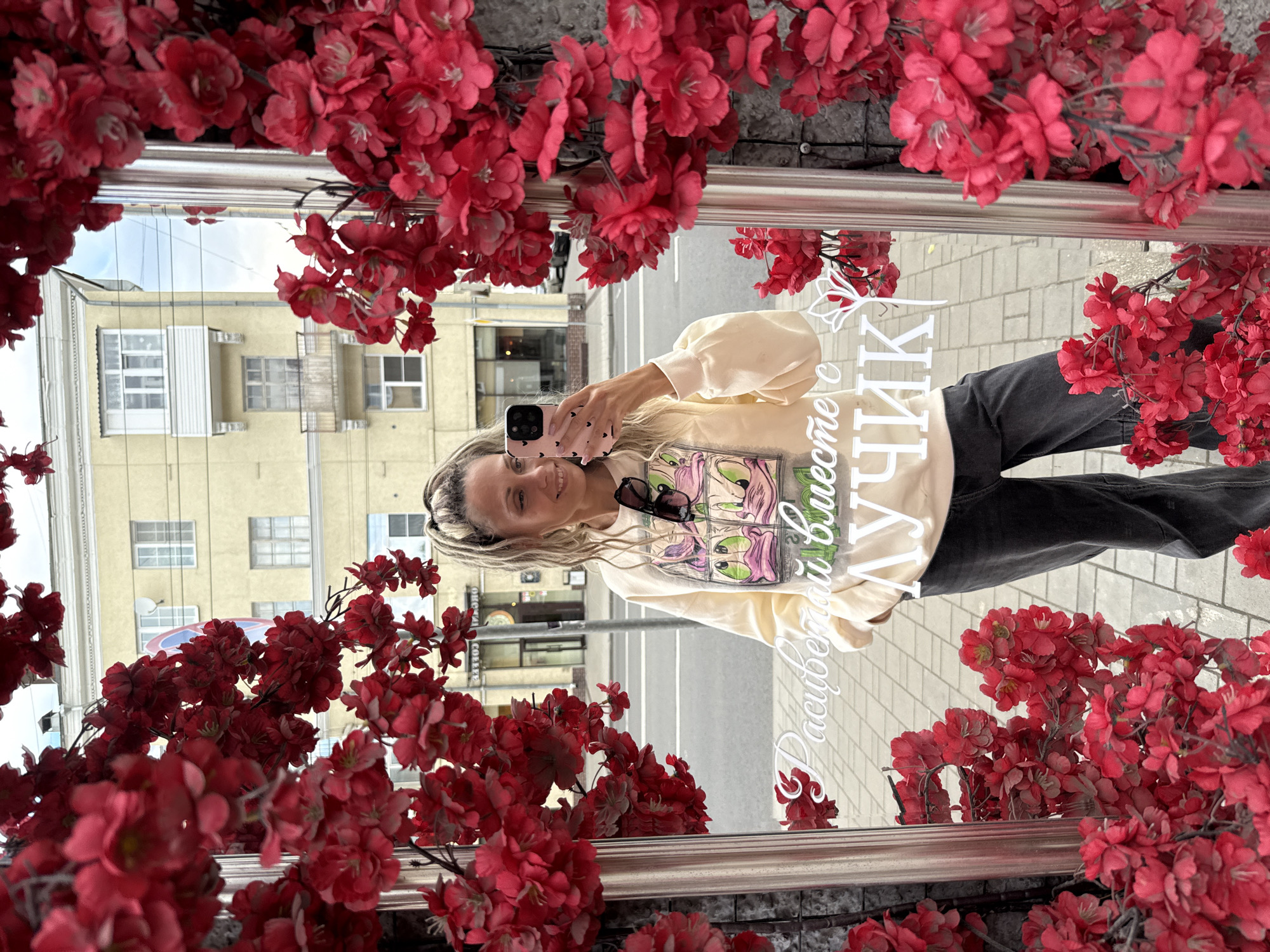 A woman with blonde hair styled in pigtails is taking a selfie in a mirror decorated with artificial red flowers. She is wearing a light-colored sweatshirt with a cartoon graphic and dark pants. She is holding a pink smartphone in her right hand. The mirror reflects a street scene with a yellow building and a cloudy sky. The text "Расцветай вместе с" is visible on the mirror in white lettering, followed by the word "ЛУЧИК" in larger white lettering. The scene appears to be set in a city.