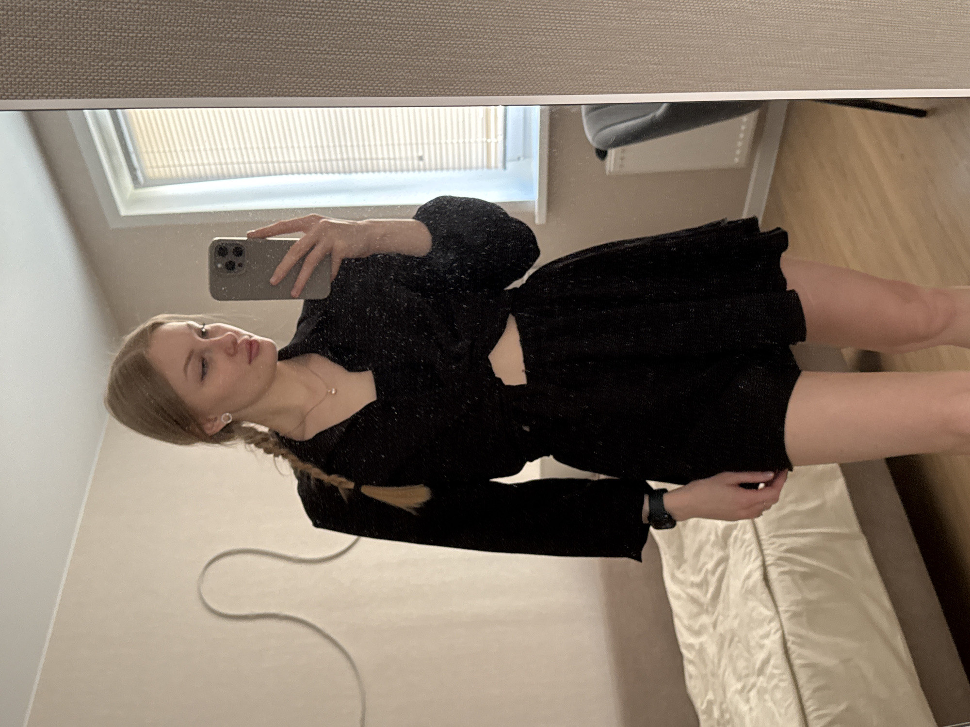 A woman takes a mirror selfie in what appears to be a domestic interior. The woman has fair skin, blonde hair in a braid, and is wearing a black outfit that appears to be a short dress or a matching top and shorts set. She is holding a smartphone in her left hand to capture the image. Visible in the background is a window with blinds, a portion of a chair, a wall with a decorative design, a bed with light-colored bedding, and a wooden floor. The location is not discernible as Perm, Russia, based on the visual elements in the image.