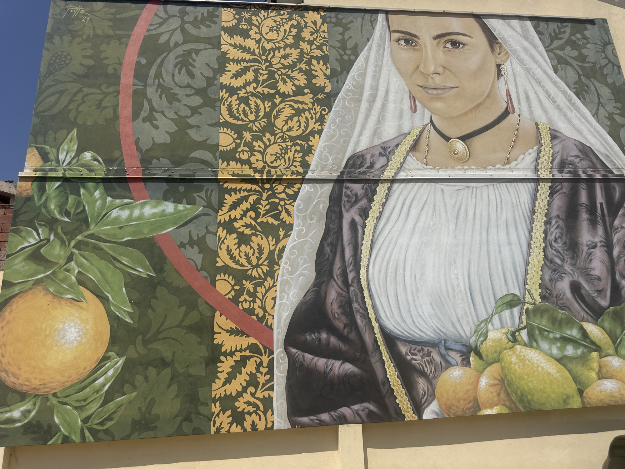 The image is a mural painted on the side of a building. It depicts a woman wearing traditional attire, including a white veil, a dark patterned dress, and elaborate jewelry. In the foreground, there are clusters of ripe oranges and lemons on branches. The mural incorporates patterned backgrounds in green and gold. To the upper left of the mural, there is a signature that appears to read "Tatte '21". The mural is located in Muravera, Italy.