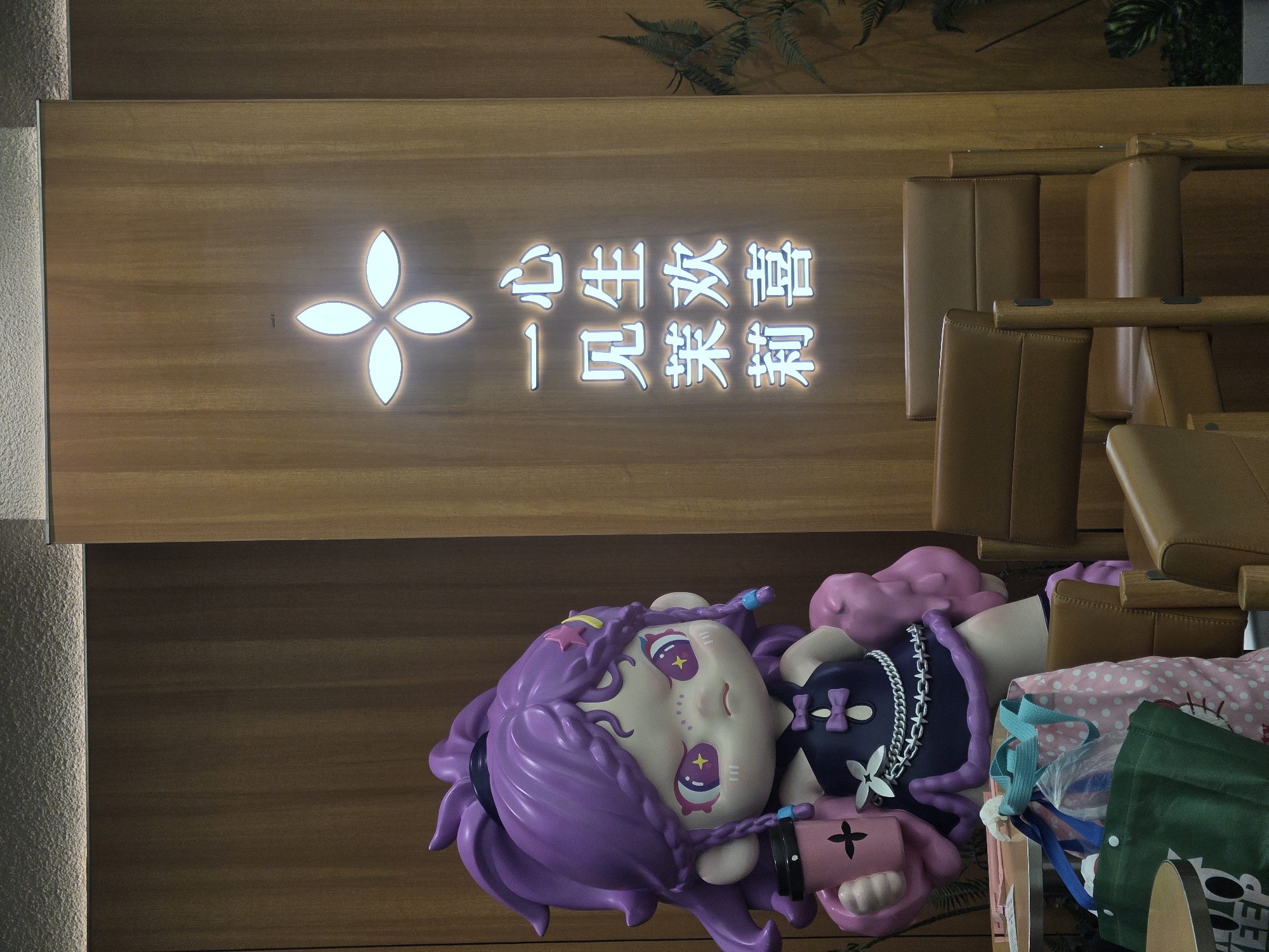 This portrait-oriented image captures an indoor scene, likely a cafe or shop in Bangkok, Thailand. A large wooden wall panel dominates the left side, featuring an illuminated white four-petal logo. Below the logo, glowing white Chinese characters are arranged vertically, reading "心生欢喜 一贝来袭." This phrase translates to "Joy arises in the heart, Yibei (a brand name) arrives/strikes," suggesting the establishment might be associated with the Yibei brand.
To the right and partially overlapping the wooden panel, a large, intricately designed figurine is prominently displayed, lying horizontally on a brown surface. The figurine depicts a character with flowing, bright purple hair styled with braids and accessories like stars. Its pale face features large, stylized eyes with striking yellow star and cross pupils, along with pink markings on its cheeks. The character wears a dark top adorned with chains and bows, and holds a pink object resembling a toy weapon or accessory.
Beside the figurine, several shopping bags are visible, including a green reusable bag and a pink polka-dotted bag, suggesting recent purchases. In the upper right corner, parts of brown leather-padded seating are visible, indicating a waiting or dining area. Subtle artificial green foliage can be seen at the very top edge of the wooden panel. The scene is brightly lit, suggesting either daytime or a well-lit interior.