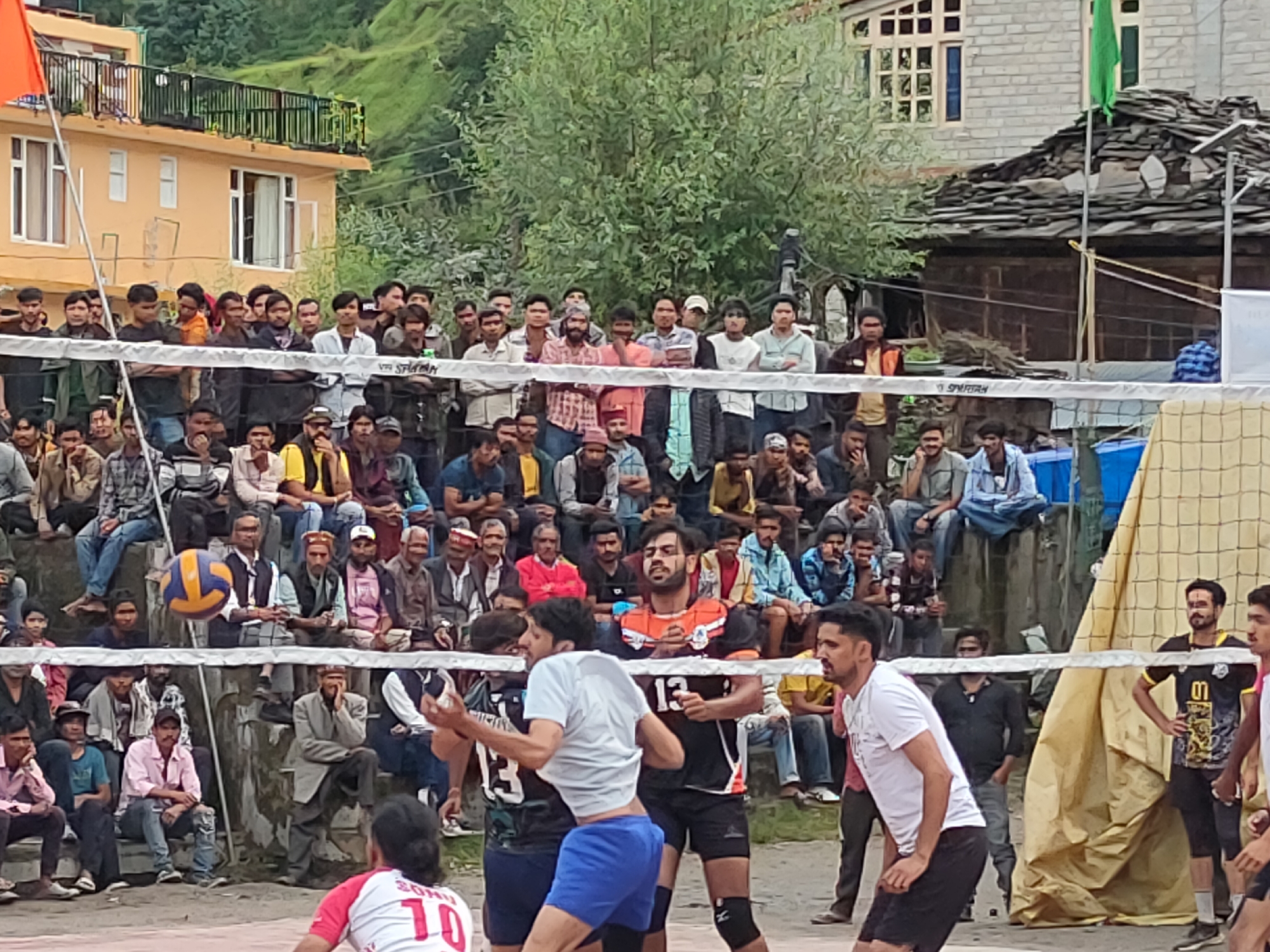 This is a photograph of a volleyball game taking place in Burwa, India. The game is being played outdoors on a dirt court with a net stretched across the middle. There are spectators gathered on both sides of the court, many of whom are sitting on tiered seating. The players are wearing jerseys and shorts, and one player in the foreground is wearing a white shirt and blue shorts. The ball is in the air, and the players appear to be in the middle of a play. In the background, there are buildings and trees, with hills in the distance. The overall scene suggests a local community event, likely a tournament or friendly match.