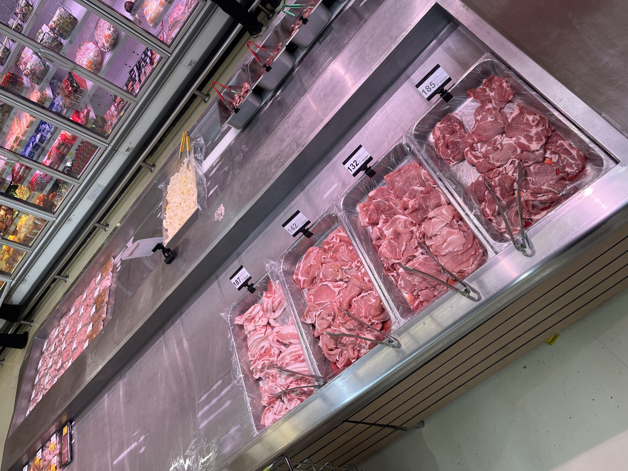 This image captures a section of a supermarket meat display in Nong Pa Khrang, Thailand. The primary focus is on trays of raw meat, predominantly pork, arranged in stainless steel containers. The meat varies in cut and marbling, with some appearing thinly sliced and others in larger pieces. Several trays are equipped with metal tongs for customer use.
In the background, a refrigerated display case is visible, filled with various packaged food items, some of which appear to be seafood or processed meats. The flooring is a light-colored, speckled surface. The overall environment suggests a well-lit, commercial retail setting. No individuals are clearly depicted in a manner that allows for detailed identification or action description. The scene is static and observational.