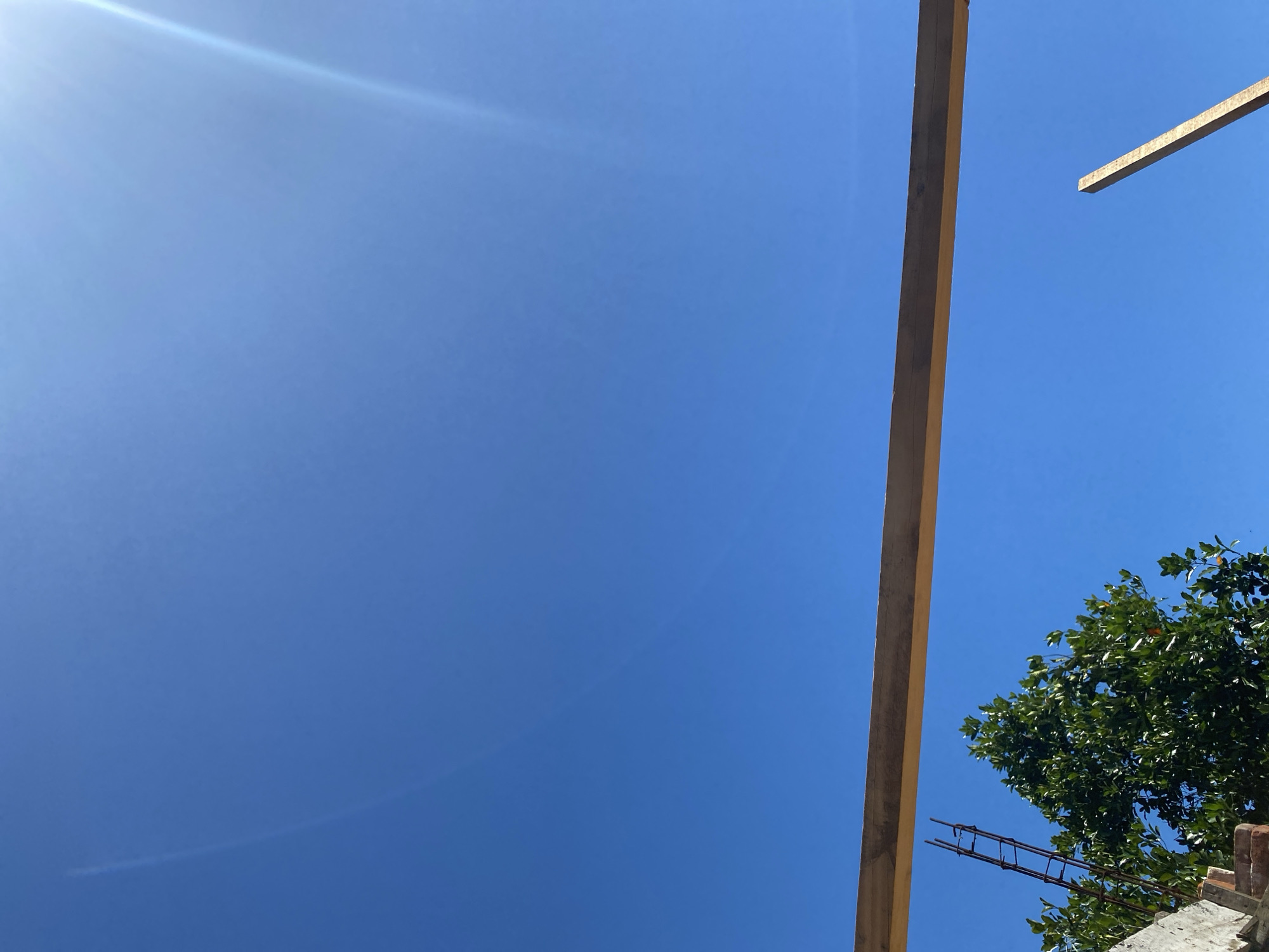 A low-angle outdoor shot captures a bright blue sky with a subtle lens flare in the upper right quadrant. In the foreground, a thick wooden beam stretches horizontally across the frame, positioned slightly above the center. Below the beam, a lush green tree with dense foliage occupies the left half of the image. To the left of the tree, a section of a brick structure is visible, and a rusty metal rebar cage rises vertically behind it. In the lower right, a thinner wooden plank is angled upwards against the sky. The scene appears to be a construction site under a clear, sunny sky.
