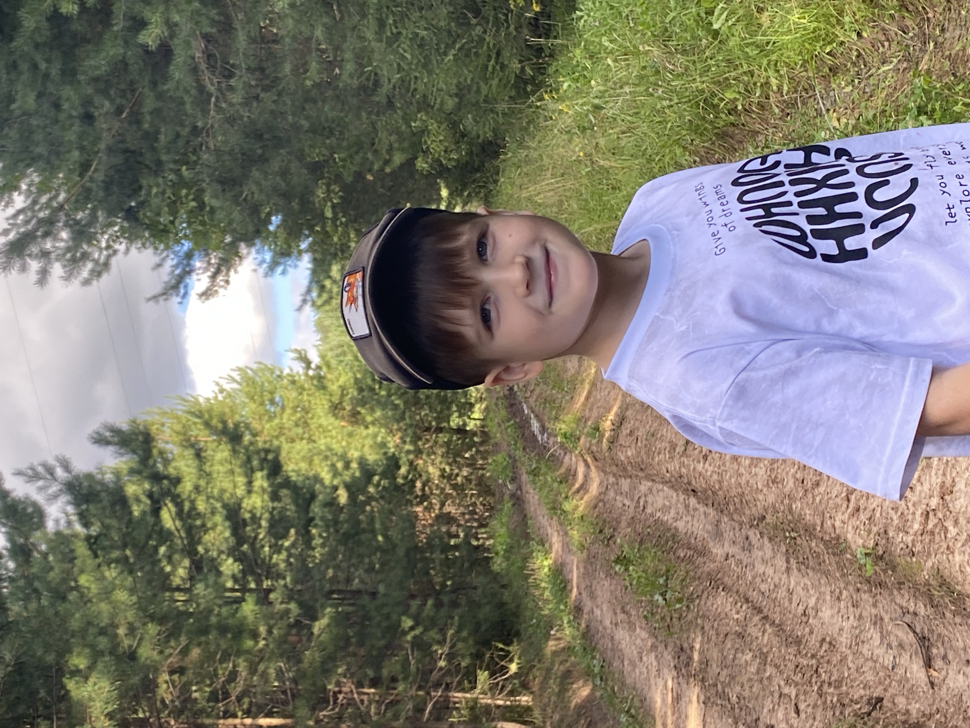 A young boy with blue eyes and brown hair is wearing a white t-shirt and a dark baseball cap with an embroidered fox on it. He is standing in a forest, on a dirt road that appears to be a trail. The trees are tall and green, and the sky is cloudy. The boy is smiling and looking at the camera. The location is a forest road in Fokino, Russia.