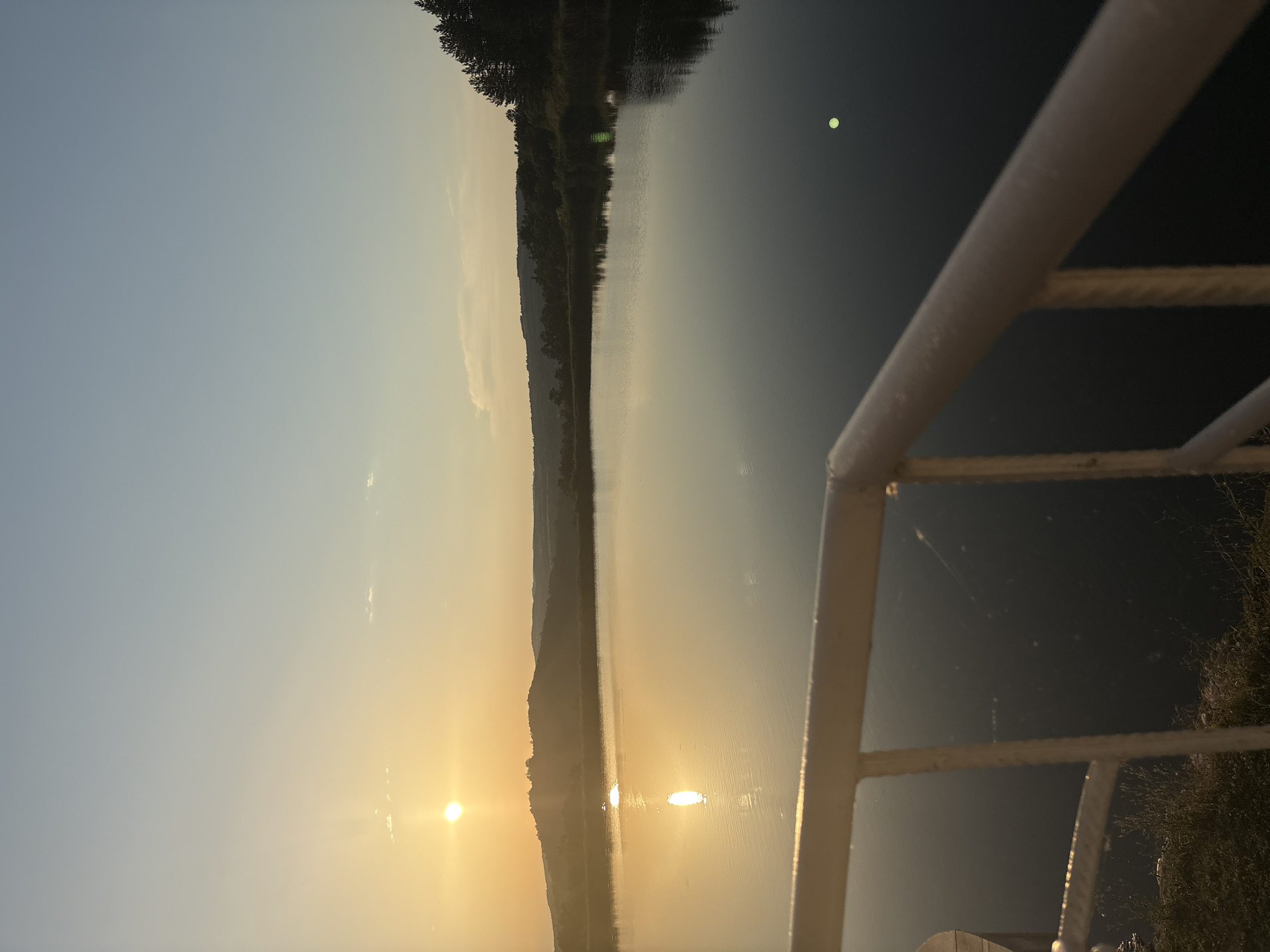 A wide, still body of water reflects the warm glow of a setting sun. The sky above transitions from a pale yellow near the horizon to a soft blue higher up. In the distance, rolling hills are silhouetted against the light, with a line of dark trees along their crest. In the foreground, a white railing, likely part of a balcony or deck, partially obstructs the view of the water. The sun, a bright orb, casts a shimmering reflection on the water's surface. The overall scene evokes a sense of tranquility and natural beauty, possibly at dusk. The location is identified as Skalsko, Bulgaria.