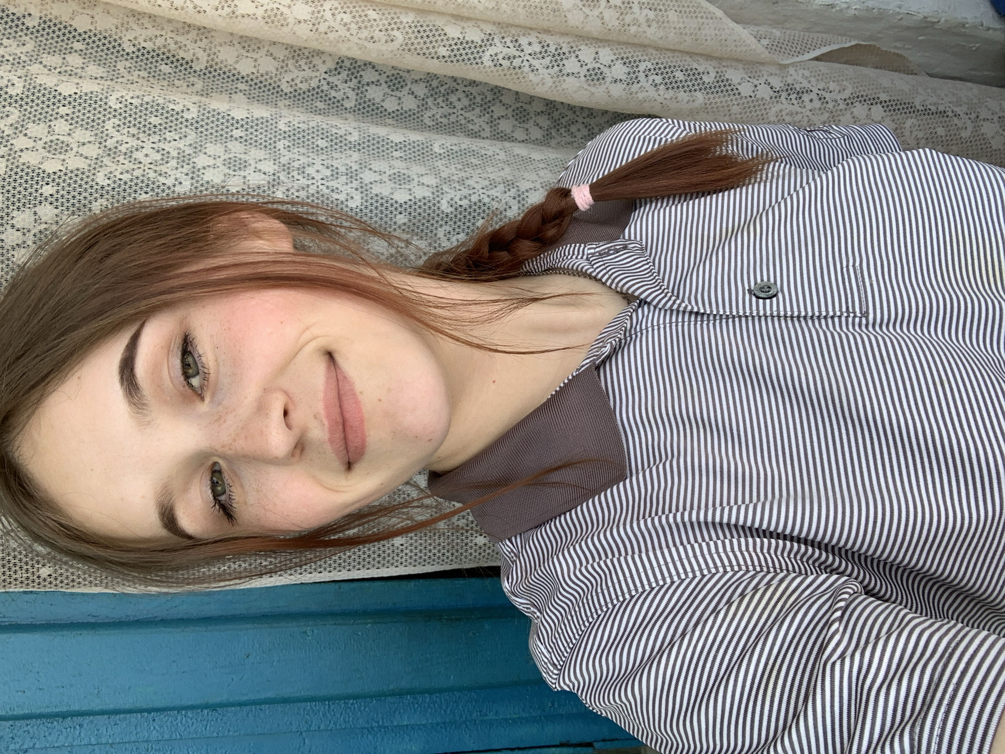A young woman with light brown hair and freckles is the primary subject. She has one side of her hair braided with a pink hair tie at the end. She is wearing a gray and white striped shirt with a dark brown undershirt. The background includes a white lace curtain and a blue surface, possibly a wall or ledge. The scene is indoors, based on the curtain and lighting. The location in the city of Фоки, Russia cannot be confirmed by this image alone.
