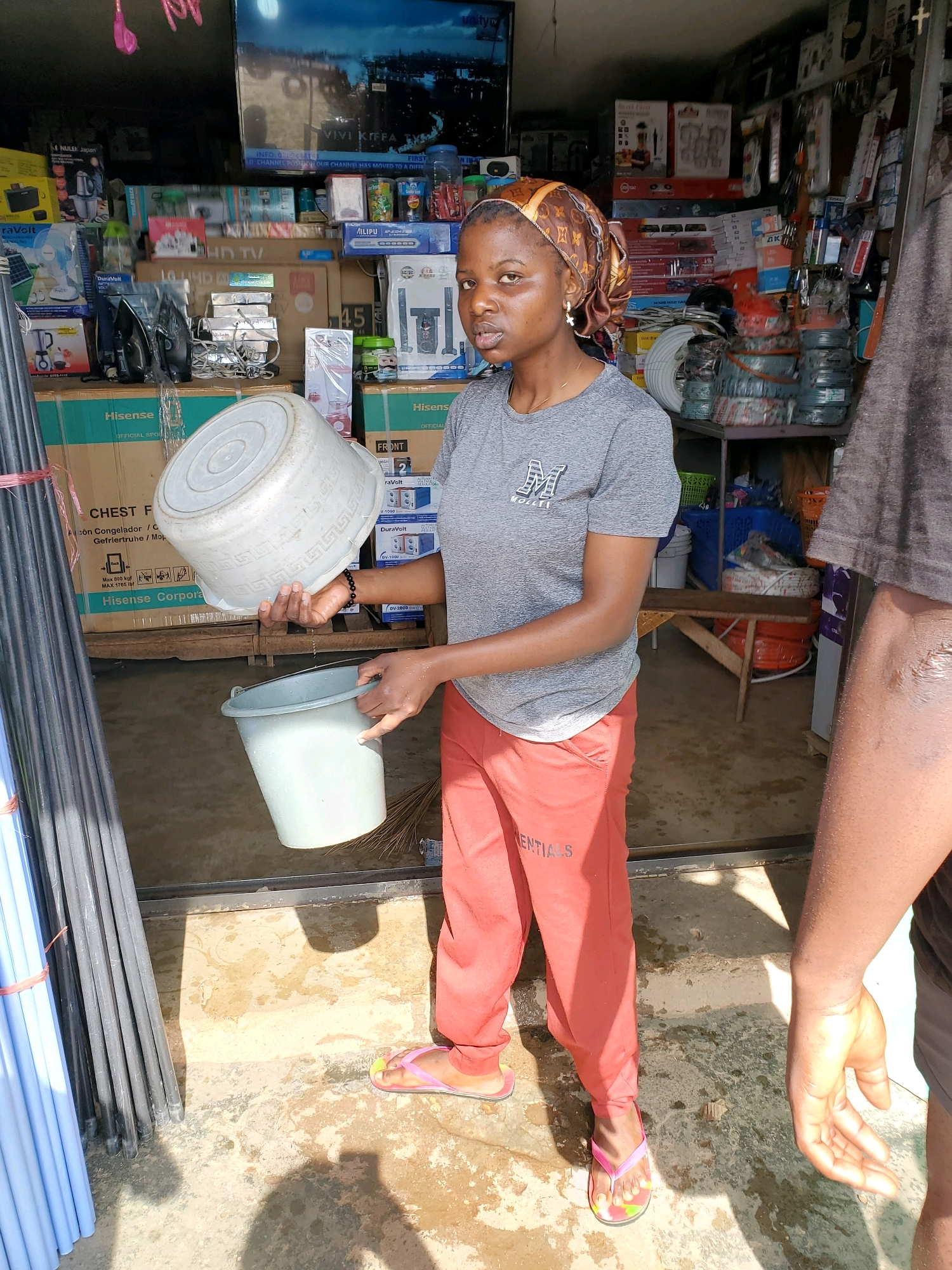 In Gura Top, Nigeria, a young woman wearing a grey t-shirt, red pants, and a headscarf is captured in a store that appears to be selling electronics and household goods. She is holding a large, white plastic bucket over a smaller, grey plastic bucket, from which water is dripping. The store's interior is visible behind her, with shelves stocked with various products, including appliances, boxed electronics, and coiled wires. The floor is a mix of concrete and dirt, with some damp areas suggesting recent activity. Another person's arm and hand are visible in the right foreground of the image.