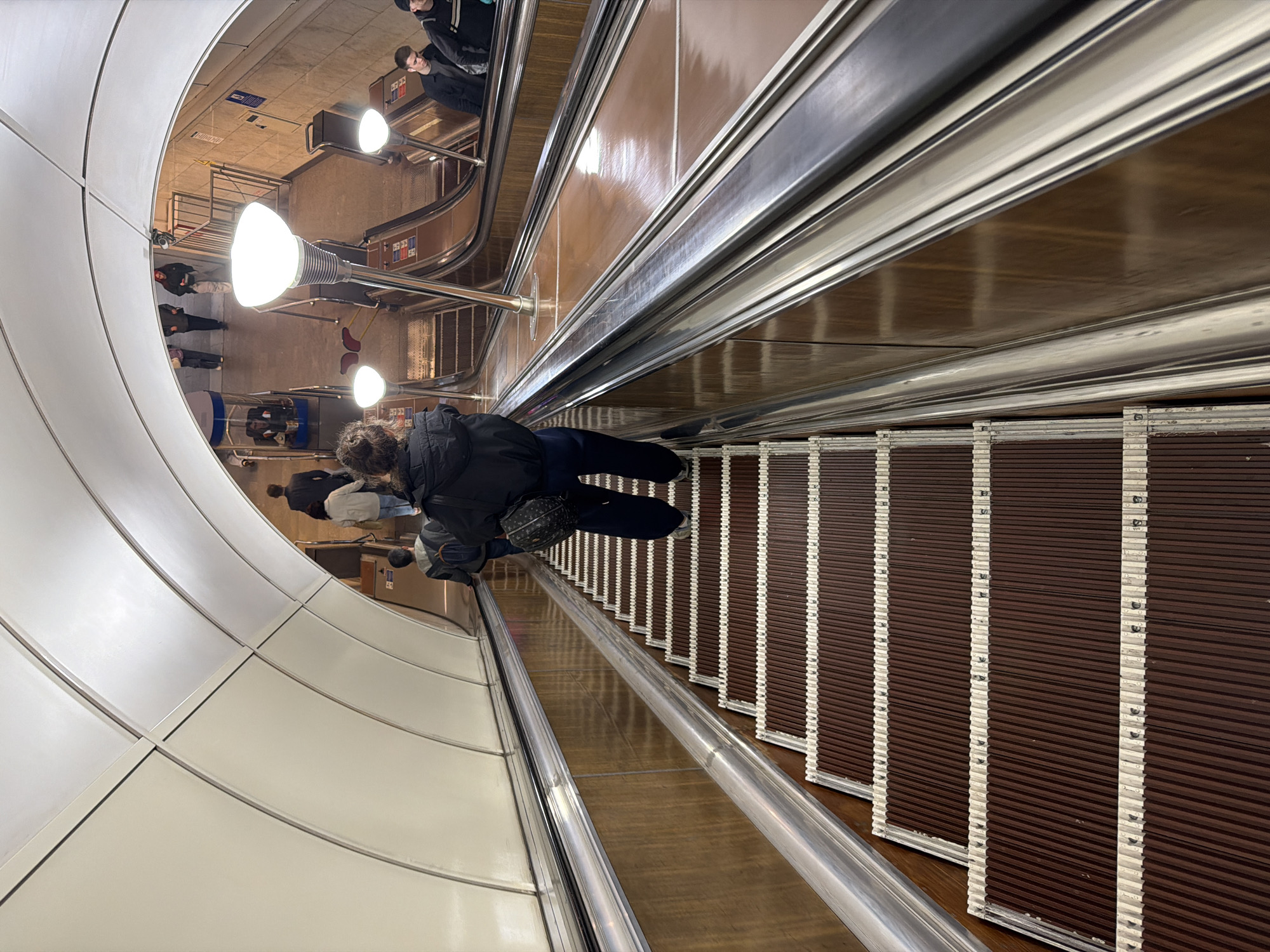 The image depicts the interior of the Avtovo metro station in St. Petersburg, Russia. An escalator is visible, with a person riding down. The station's architectural features, including the curved white walls and lighting fixtures, are prominent. Other people are seen on the station platform.