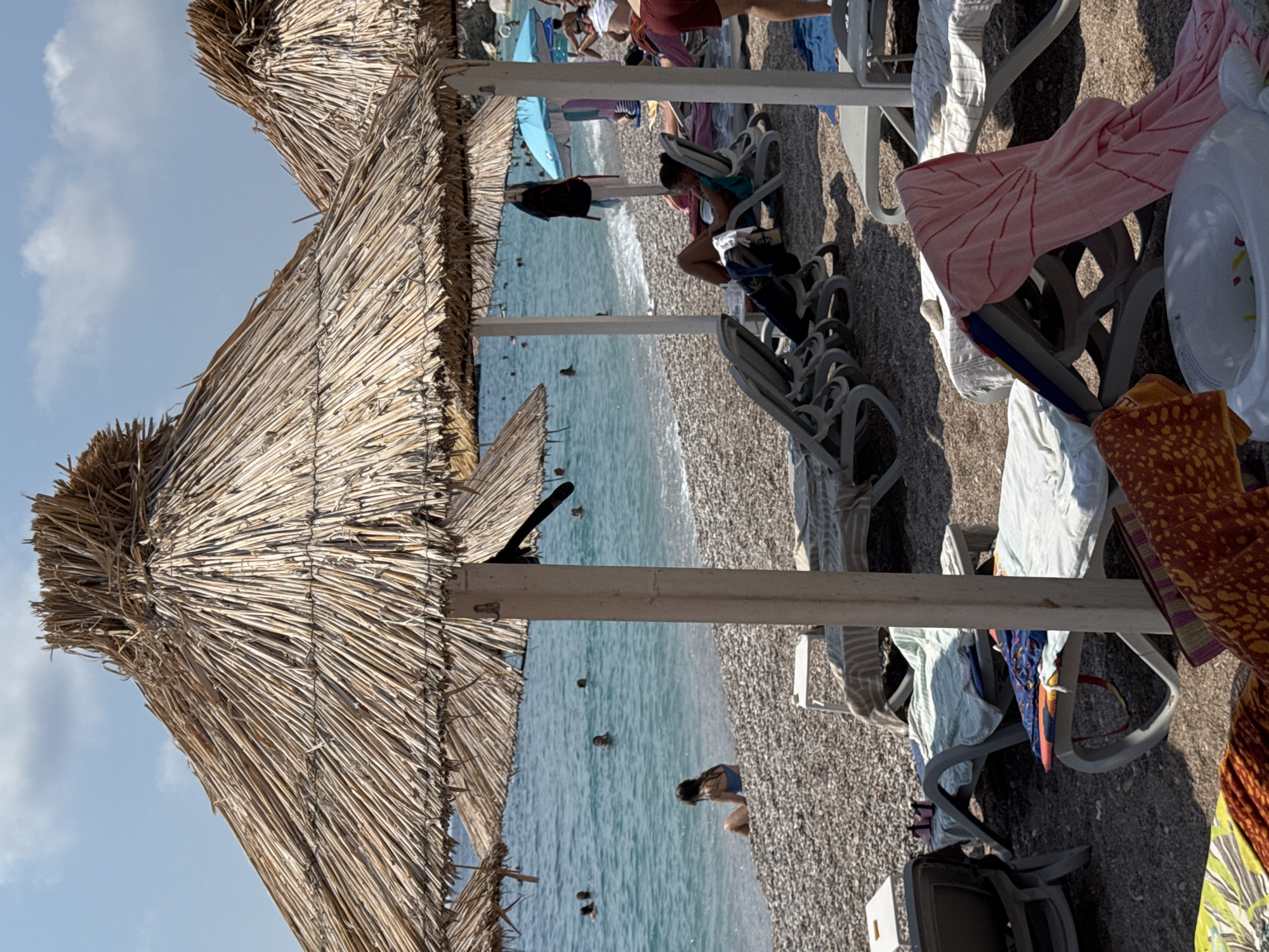 The image depicts a crowded beach scene in Mirište, Montenegro, under a clear blue sky with scattered clouds. The foreground is dominated by several straw-thatched beach umbrellas, casting shadows on the pebble-covered shore. Beach chairs are arranged beneath the umbrellas, some occupied and others empty, with towels and blankets draped over them.
In the middle ground, the turquoise waters of the Adriatic Sea are visible, with several people swimming and wading near the shore. A woman in a blue swimsuit sits on the pebbles, facing the water. Further down the beach, more umbrellas and beachgoers can be seen.
The environment is characterized by the pebble beach, the calm sea, and the presence of beach amenities like umbrellas and chairs, indicating a typical summer day at a popular coastal resort. The overall impression is one of a vibrant and well-frequented tourist destination.