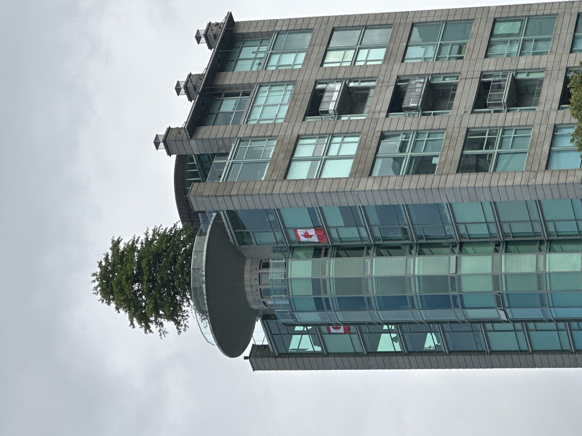 A high-angle, close-up, full-frame, eye-level outdoor shot captures the upper portion of a modern, mixed-use building in Vancouver, Canada, against a heavily overcast sky. The building features a distinctive cylindrical glass section on its left side, adorned with two small Canadian flags. To the right of this cylindrical section, the building presents a more rectangular facade, characterized by rows of large, square, light-blue tinted windows and balconies with glass railings. The building's exterior is a combination of stone cladding and extensive glazing. At the very top of the cylindrical section, a large, circular balcony with a glass railing is visible, supporting a mature, lush green tree. The overall atmosphere is muted due to the cloudy weather.