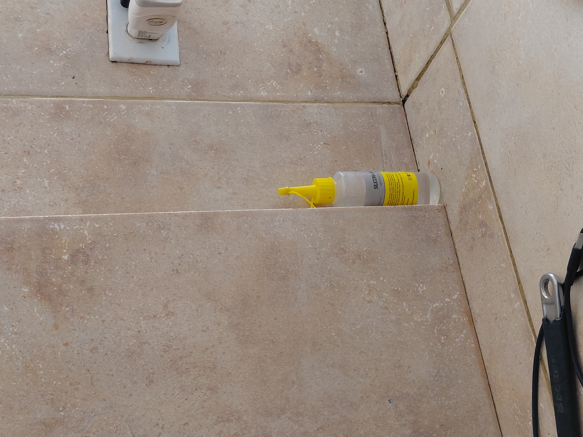 This image displays a corner of an indoor tiled surface, likely a floor or a low wall, in Santa Ana, Venezuela. The tiles are light brown or beige, featuring a speckled, mottled pattern, and are separated by grout lines.
In the upper left quadrant, a white electrical outlet is visible, with a light-colored charger plugged into it. The charger has a logo that appears to read "Dinsey" and includes small regulatory symbols like a CE mark and a crossed-out wheeled bin.
Centered in the image, resting on a tile ledge or step, is a small, clear plastic bottle with a bright yellow screw-top cap and a pointed nozzle. The label on the bottle is yellow, and the most prominent visible text reads "SILICONA U". Below this, "USO GE..." is partially visible, indicating it's likely a universal silicone sealant or adhesive. The bottle contains a clear liquid and appears to be about two-thirds full.
To the far right, a black strap or cable with a metallic loop attachment is partially visible, extending into the frame from the right edge. The grout lines between the tiles are visible, and in some areas, there appears to be fresh sealant or a similar material applied along the seams.
No specific activity is occurring; the objects are static. The lighting suggests an indoor setting, and no time of day or weather conditions can be determined.