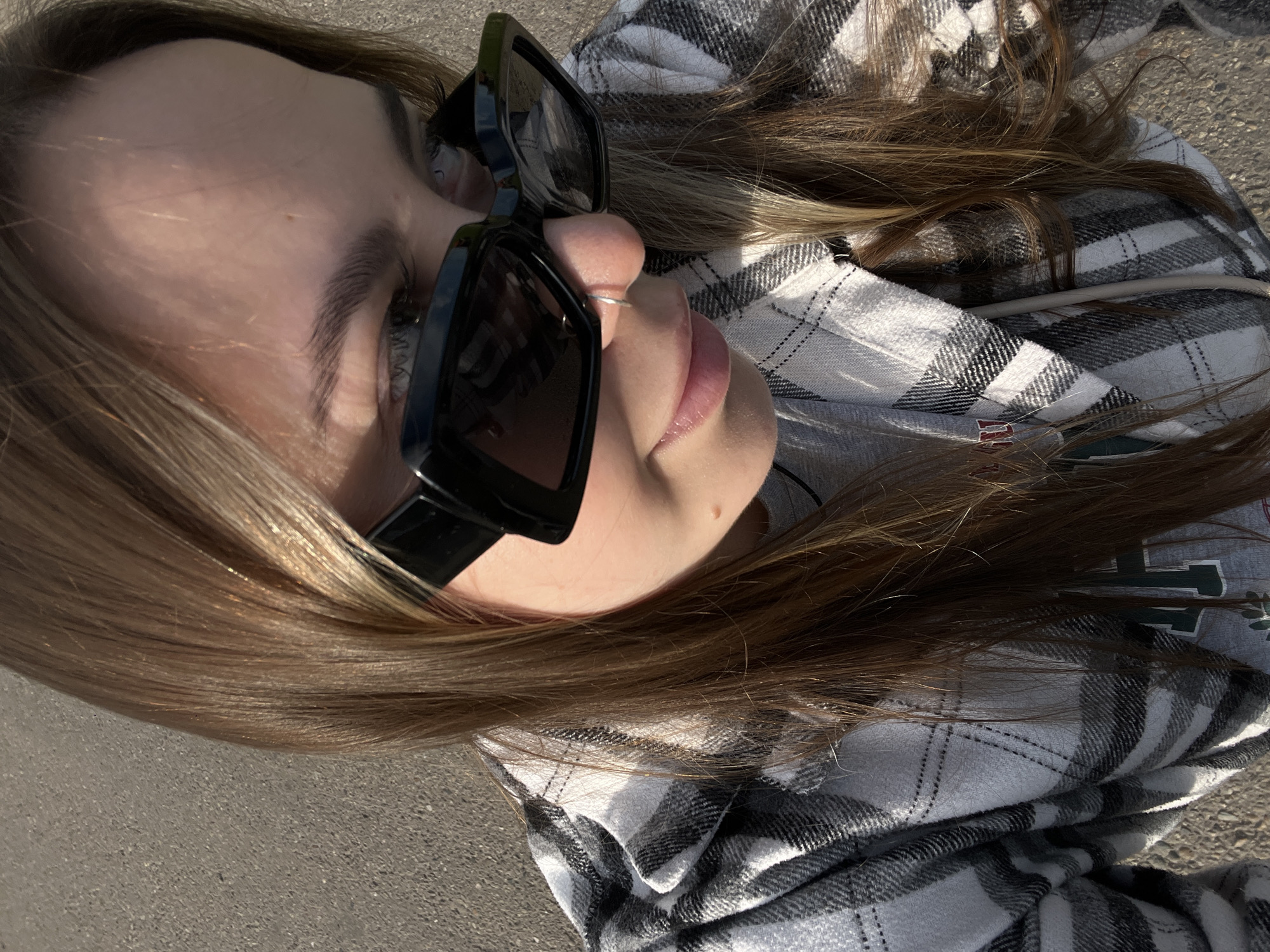 A young individual, appearing female, is captured from a high-angle perspective, looking downwards and slightly to their right, with their head tilted. They have long, light brown hair with sunlit highlights. The individual is wearing thick, dark eyebrows, and a small silver hoop nose ring is visible in their left nostril. A subtle smile is present on their lips. They are wearing large, black, rectangular-framed sunglasses. The individual is dressed in a grey undershirt with a partially visible green and white design on the left side, covered by a black, white, and grey plaid long-sleeved shirt or jacket. A small mole or beauty mark is visible on the right side of their neck/collarbone area. The background consists of a textured grey surface, consistent with asphalt or concrete pavement, indicating an outdoor setting in the city of Novopokrovskaya, Russia. Direct sunlight casts strong highlights on the hair and clothing, suggesting a bright daytime environment.