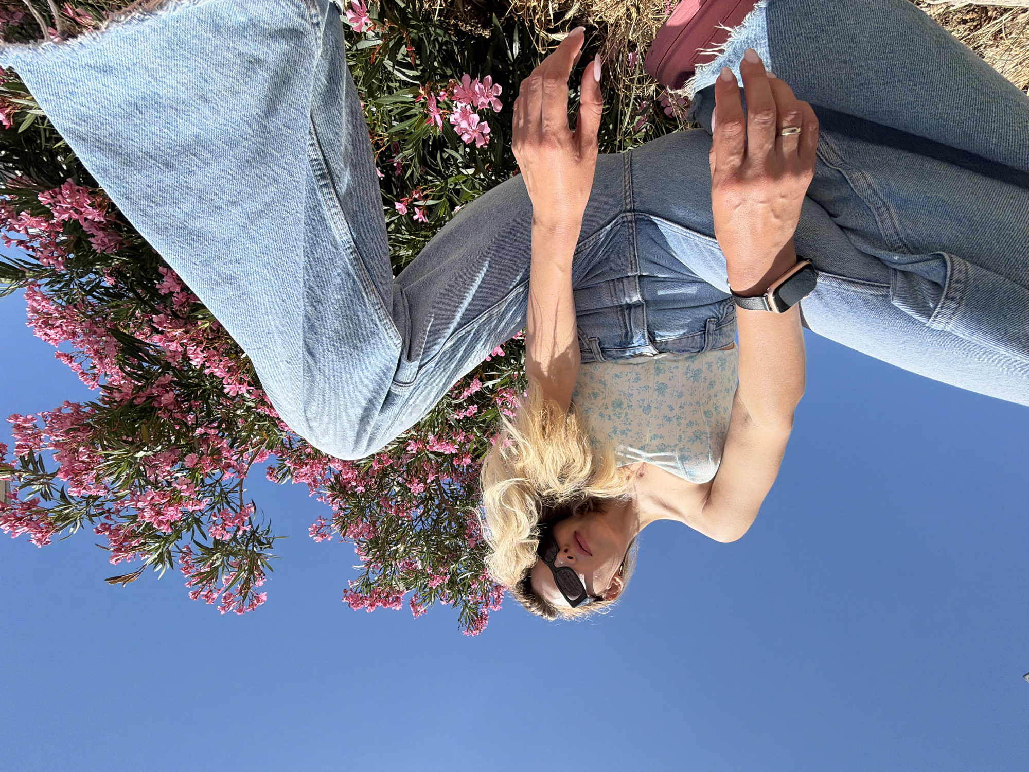 An upward-angled outdoor photograph, taken in Budva, Montenegro, captures an individual seated or crouching against a clear blue sky. The person, with long blonde hair cascading over their right shoulder, wears black sunglasses, a light blue and white floral strapless top, and light-wash, wide-leg denim jeans with frayed hems. A smart watch featuring a rose gold case and black band is present on the left wrist, and a plain silver-toned ring is worn on the left ring finger. The individual's head is tilted back and slightly to the right, with their gaze directed downward. To the right of the frame and extending behind the individual is a large oleander shrub, dense with clusters of pink flowers and green foliage. The immediate foreground beneath the person consists of dry, light-colored earth with scattered pink petals. A dark, thin vertical object is faintly visible against the sky in the upper left quadrant.