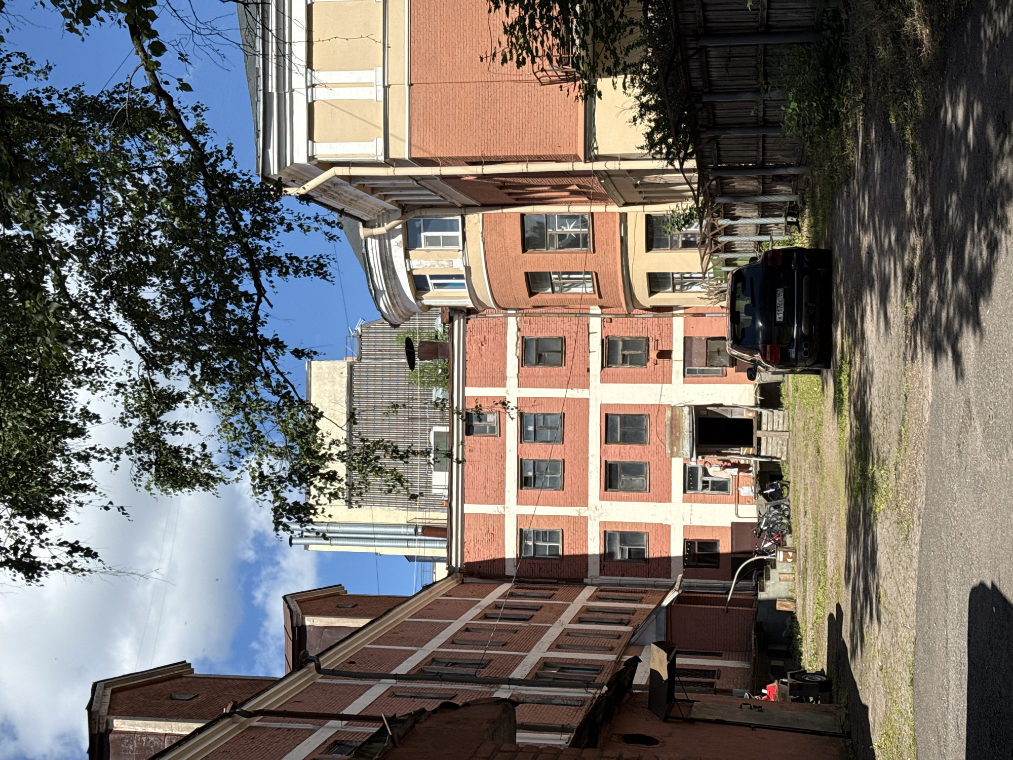 The image depicts a courtyard area in Vyborg, Russia, enclosed by brick buildings. The buildings, primarily constructed of red brick with white architectural details, appear to be multi-story structures with numerous windows. In the foreground, a paved pathway leads into the courtyard, where a black SUV is parked. Adjacent to the pathway, a wooden fence with overgrown vegetation runs along the right side of the image. Further into the courtyard, a person is visible standing on a porch area near an open doorway. Several bicycles are parked near the porch, and a red trailer with a wheel is situated on the left side of the frame. Overhead, tree branches with green leaves cast shadows across the scene. The sky above is blue with scattered white clouds.
