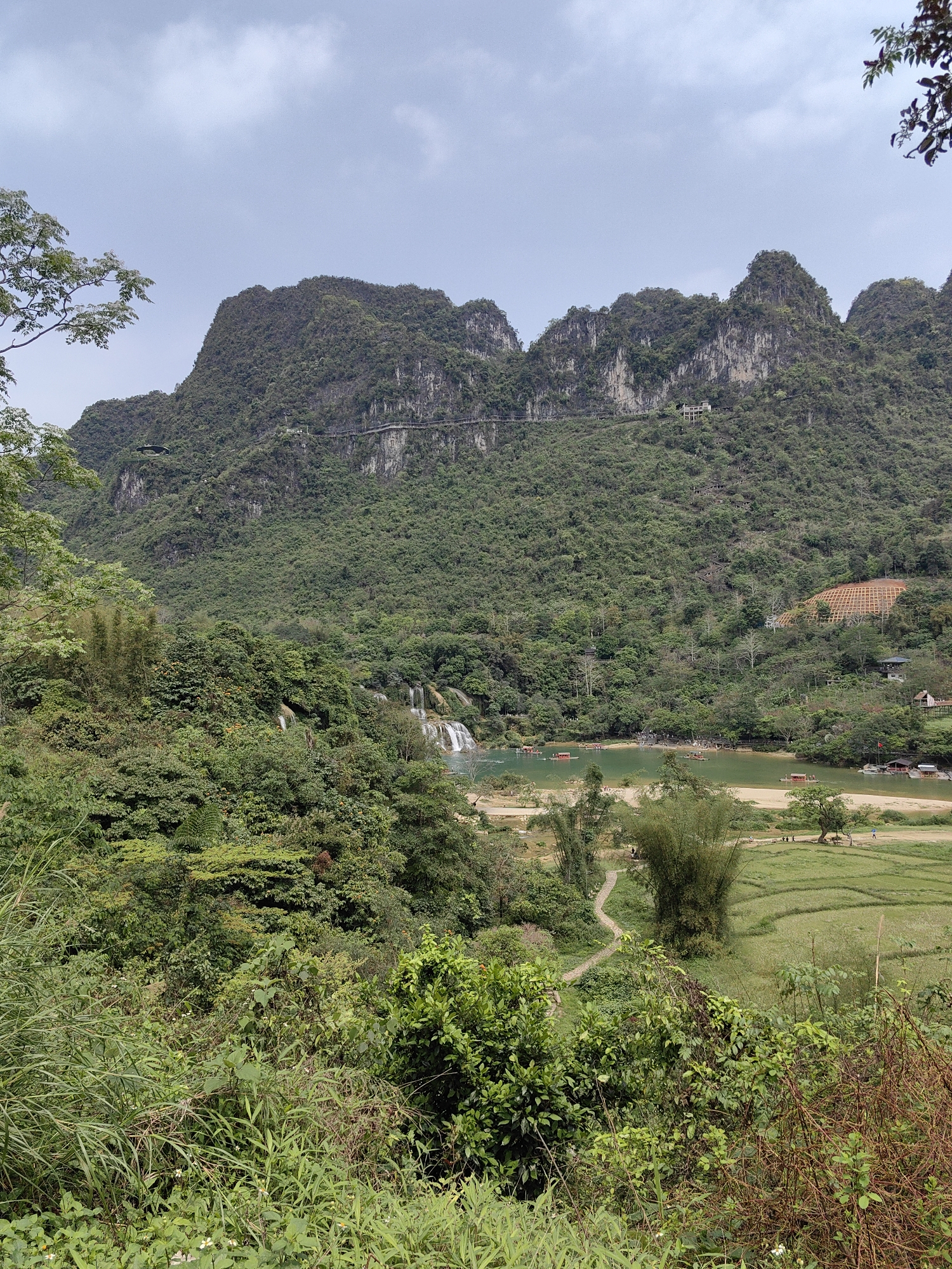 The image shows a landscape in Daxin County, China. In the foreground, there's lush greenery, with diverse foliage including shrubs, grasses, and bamboo. A narrow, dirt path winds its way through this vegetation.
Behind the foreground, there is a clear, shallow body of water which features small waterfalls cascading into it. There are several red boats are visible on the water.
Beyond the water is a valley filled with terraced rice paddies and some sparse tree cover. On a nearby hillside, some residential buildings can be seen.
The background features a mountain range covered with dense, green vegetation and rocky outcroppings. A horizontal structure resembling a walkway or road has been constructed across the face of the mountain. Above the mountain, the sky is overcast with light gray clouds.