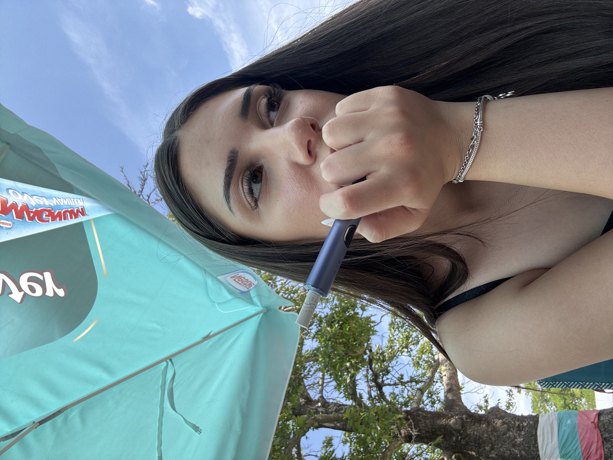 The image depicts a young adult female positioned outdoors, gazing to her right (viewer's left) with a somewhat distant expression. She has long dark hair and brown eyes, with visible facial pores and applied eye makeup. Her right hand is raised, partially covering her mouth and chin, and she holds a dark blue/purple electronic device, likely a heated tobacco or vaping product, from which a white cylindrical insert protrudes. A silver chain bracelet with multiple strands and a small charm is present on her right wrist. She is wearing a dark strap, possibly part of a top or swimsuit. Overhead, a large turquoise-colored umbrella provides shade, displaying reversed text, including "NESTLE" and "MUGAM" (likely "MAGNUM"). In the background, a large tree with green foliage is visible against a partly cloudy blue sky. Around the tree's trunk, a fabric strip displaying horizontal bands of white, green, and pink/red is wrapped. The scene is located outdoors, identified as being in Bulgaria by the national flag colors on the tree, and, as per the prompt, is specifically in the city of Kostievo.