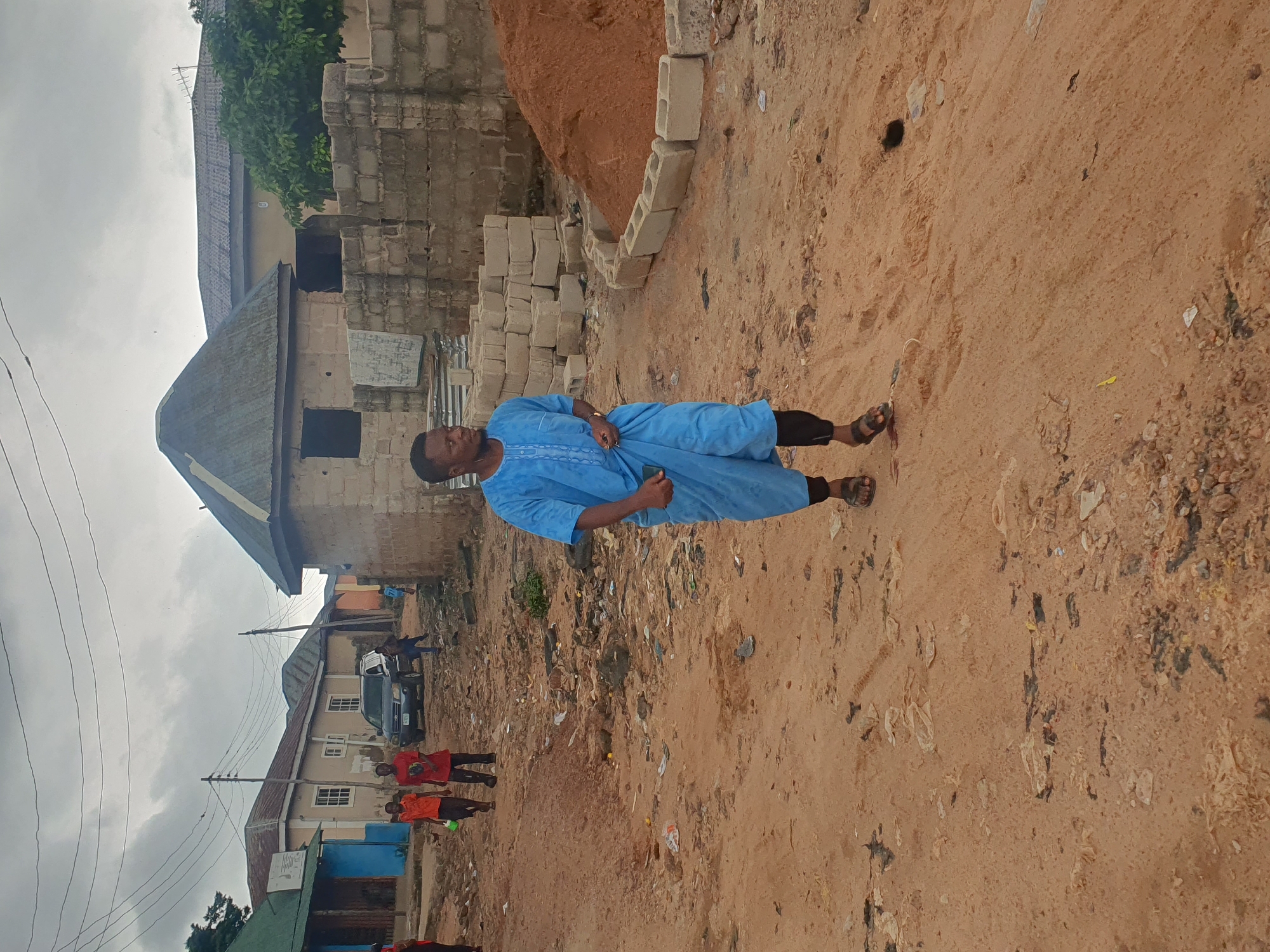 In Lokoja, Nigeria, a man in a vibrant blue traditional outfit walks on a dusty, unpaved street littered with debris. He wears black leggings and sandals. Behind him, two young boys in orange and red shirts walk in the same direction. In the background, several buildings are visible, some under construction and others appearing to be older structures. A dark-colored SUV is parked further down the street, and a pile of sand and building blocks suggests ongoing construction or renovation work. The sky is overcast, contributing to a muted lighting condition. The scene depicts a typical streetscape in a developing area, characterized by incomplete infrastructure and everyday life activities.