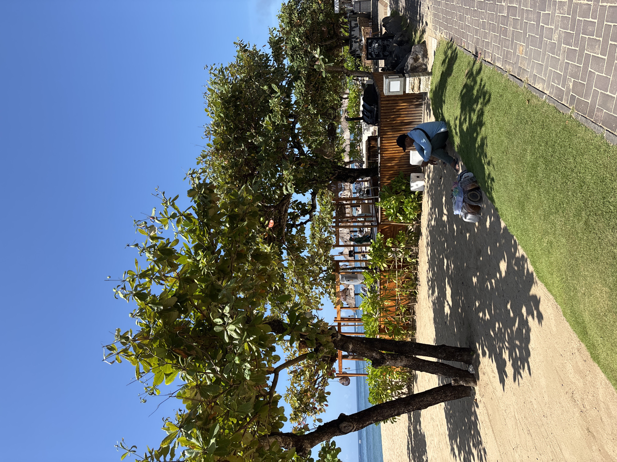 A sunny day at a beach in Kuta Selatan, Indonesia. A man in a light blue shirt and dark pants kneels on a patch of grass near a beachside path, apparently attending to belongings. A wooden structure behind him suggests a beachside establishment. Trees provide partial shade, their shadows cast on the sandy path. The ocean is visible in the background.