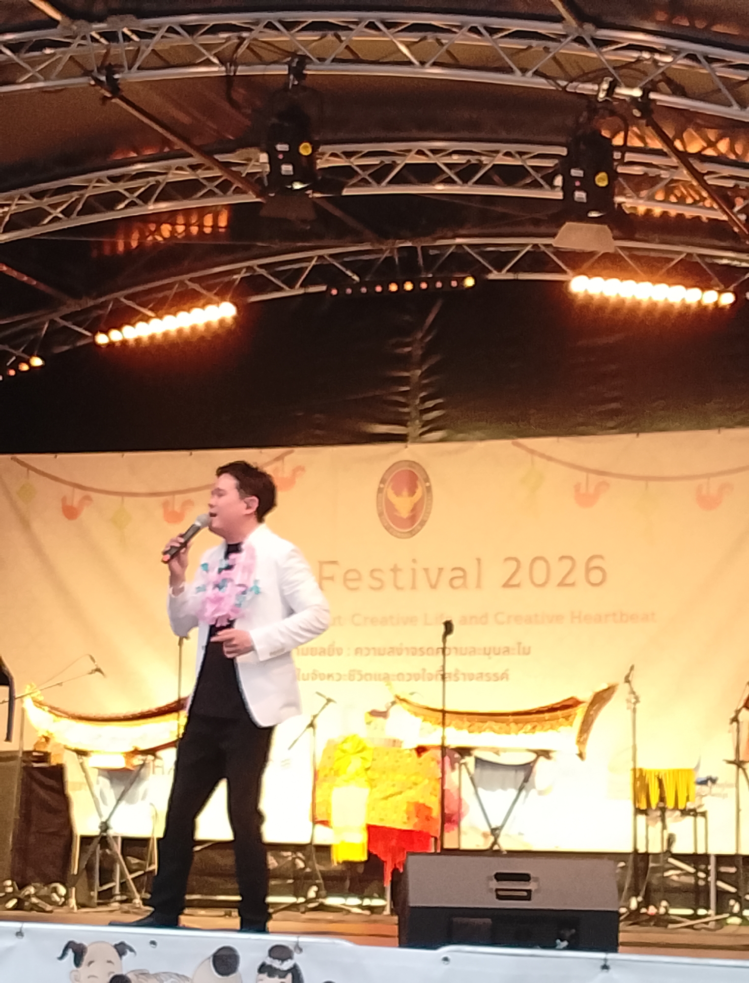 A man in a white blazer and black pants stands on a stage holding a microphone and singing. He is wearing a pink and white lei. Behind him, a banner reads "Festival 2026" in large font. The stage is lit by several rows of warm-toned lights. There are speakers and musical instruments visible on the stage, suggesting a performance is in progress. The backdrop is a light yellow with colorful decorations.