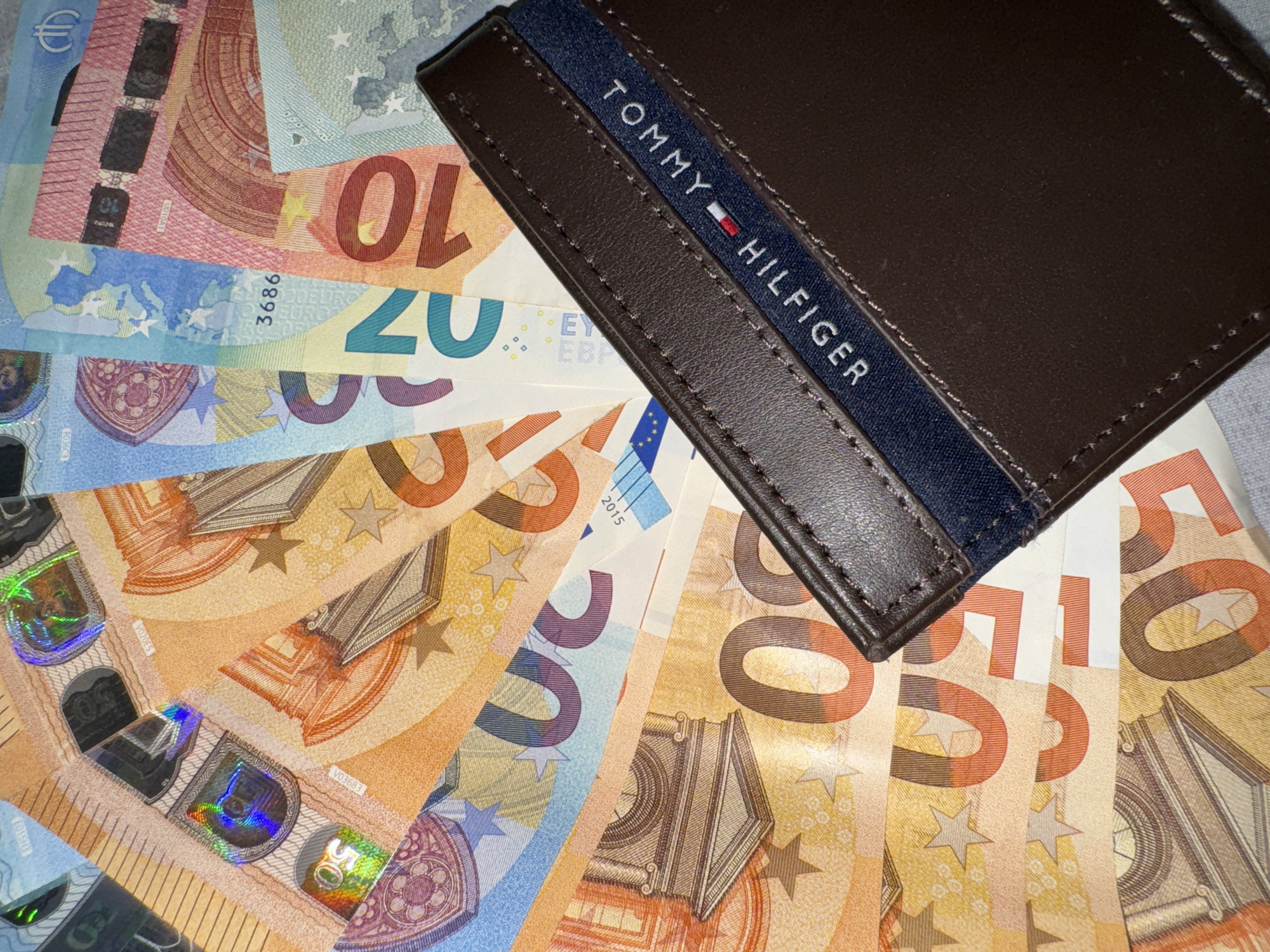 The image displays a brown Tommy Hilfiger wallet lying on top of a fan arrangement of Euro banknotes. The denominations visible include 10, 20, and 50 Euro notes. The background is a neutral color. There is no indication of location or city. No people are visible.
