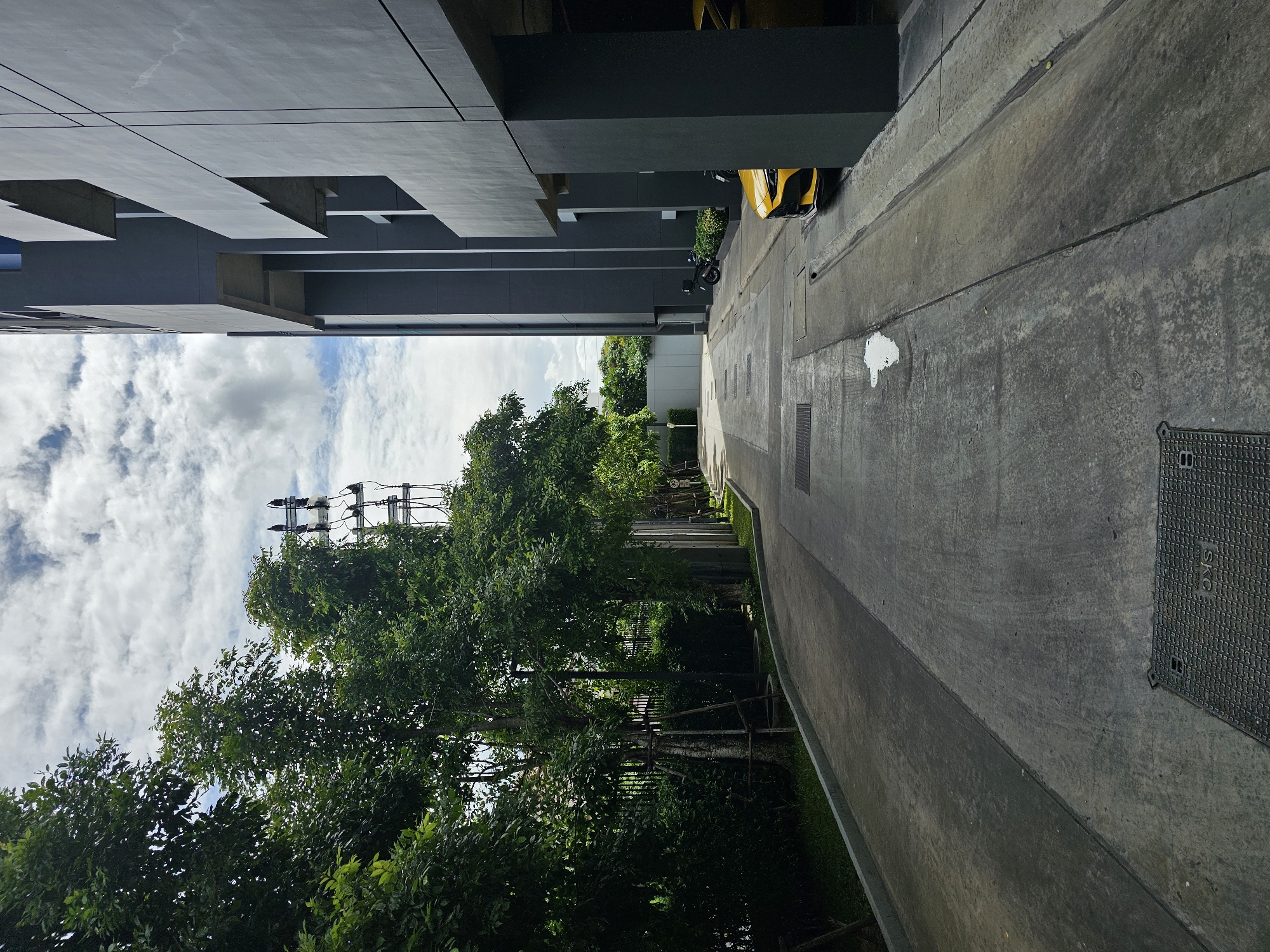 This is a high-angle, eye-level, outdoor, daytime shot taken from a slightly elevated perspective, looking down a paved driveway that curves gently to the right. The driveway is lined with trees on the left and a modern, dark grey building with protruding sections on the right. The sky is partly cloudy with white cumulus clouds against a blue background.
On the left side of the driveway, there are lush green trees and bushes, with some bare-branched trees supported by wooden stakes. A black metal fence runs behind the vegetation. Further down the driveway, a tall electrical pylon with multiple crossbars and insulators is visible against the sky.
The building on the right is a multi-story structure with a dark grey facade, featuring geometric cutouts and deep recesses. A bright yellow sports car is parked partially under the overhang of the building. Two motorcycles are also visible parked near the building's entrance.
The paved driveway itself is concrete and shows signs of wear, with some patches of dampness and a few manhole covers embedded in the surface. The overall scene appears to be a residential or commercial complex in Bangkok, Thailand, likely an apartment building or a hotel, given the modern architecture and the presence of a driveway with parking. The time of day is daytime, as indicated by the shadows and the lighting conditions. There are no people visible in the image.