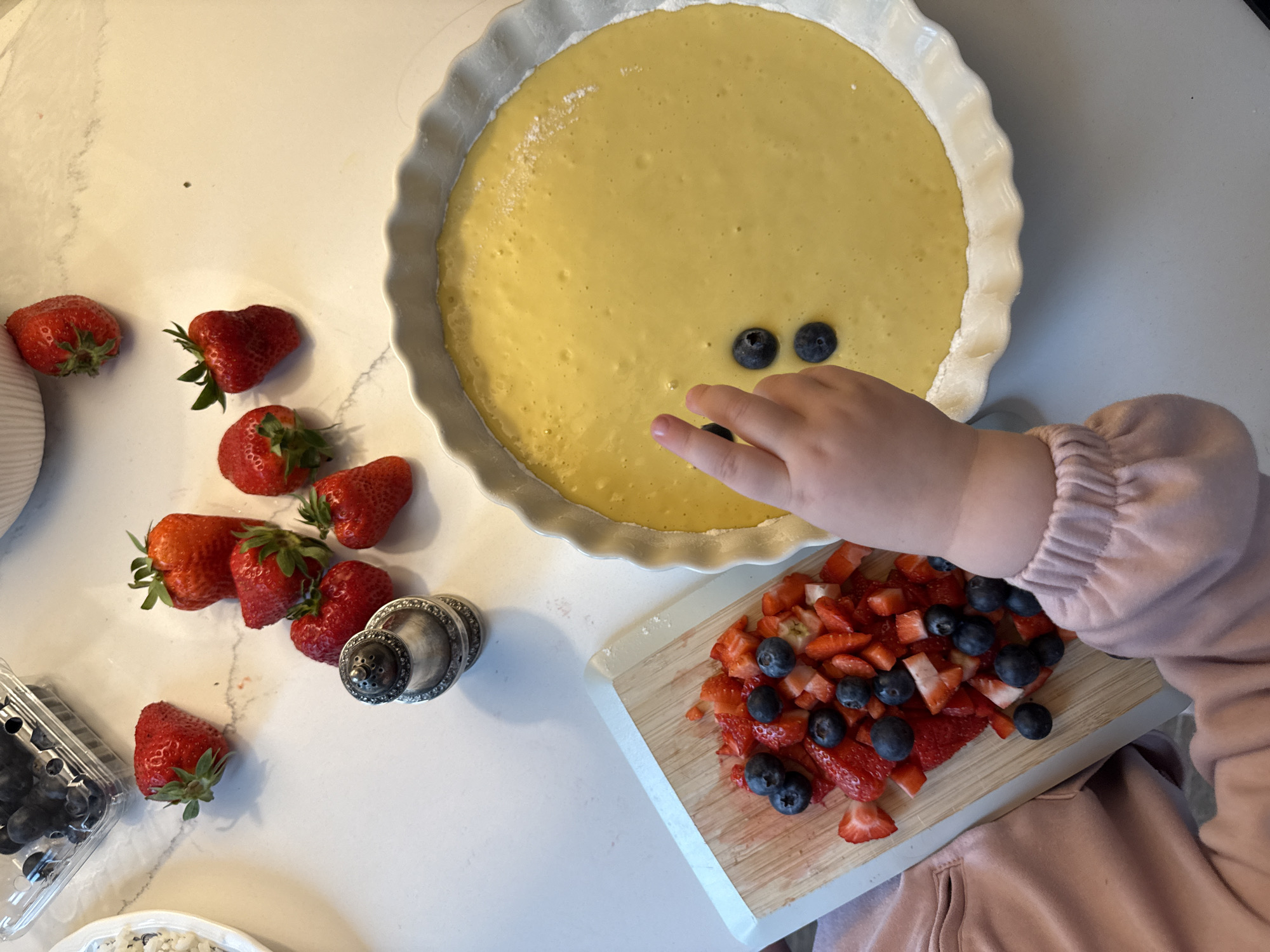 A child's hand is placing blueberries on a yellow batter filling in a white, fluted tart pan on a white countertop. A wooden cutting board with chopped strawberries and blueberries is to the right of the tart pan. To the left of the pan are several whole strawberries, a container of blueberries, and a silver sugar shaker. The child is wearing a pink sweater. The scene is likely a home kitchen. The image does not contain information about the city of Moscow, Russia.