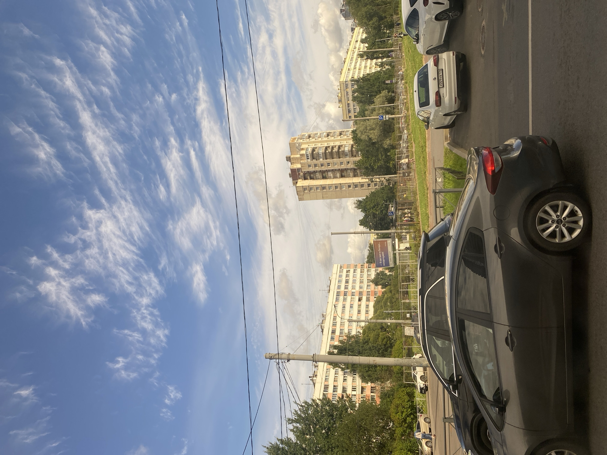 The image depicts an urban scene in Saint Petersburg, Russia, characterized by a clear blue sky with wispy cirrus clouds, suggesting fair weather. Several residential buildings, typical of Soviet-era architecture, dominate the background. These multi-story buildings feature a light-colored facade with numerous windows and balconies.
In the foreground and middle ground, various cars are visible. A dark-colored sedan is prominently parked on the right side of the road, with its rear facing the camera. A white sedan is parked next to it. Further left, a dark SUV is parked, partially obscuring a light-colored sedan in front of it. Additional cars and a black SUV are seen in the middle ground, along with parked vehicles in a lot behind a fence.
A paved road occupies the bottom portion of the image, with white lane markings. A grassy strip with a fence runs between the road and the parking area. Several utility poles with numerous wires are scattered throughout the scene, indicating the presence of electrical and communication infrastructure. Green trees and bushes are interspersed between the buildings and along the roadside, adding a touch of nature to the urban environment.
A billboard with text and an image is visible on the right side of the frame, suggesting commercial activity. Several individuals can be seen walking in the background, contributing to the sense of an active urban space. The overall impression is a typical street view in a residential area of Saint Petersburg.