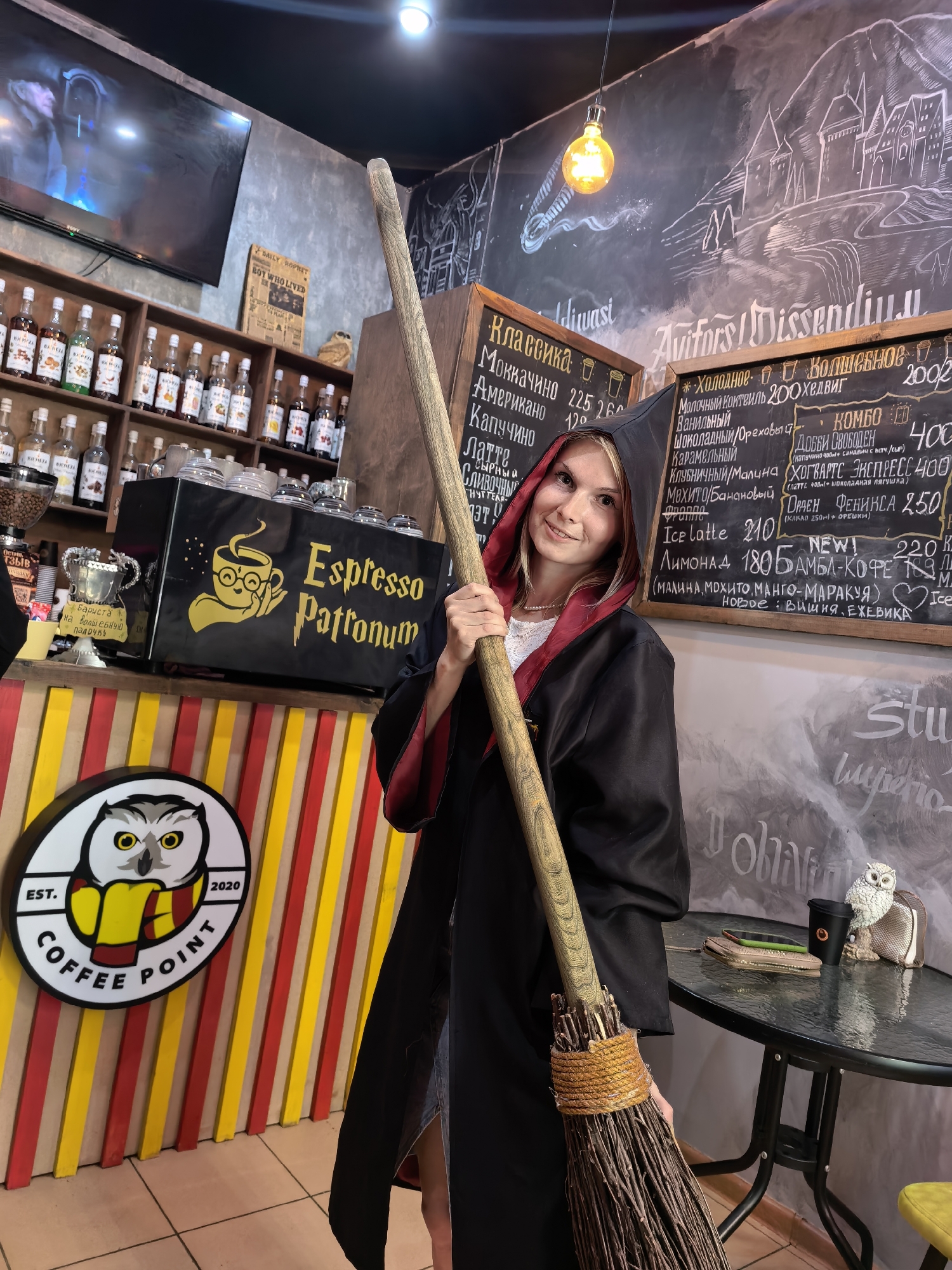 A young woman dressed as a witch stands in a coffee shop in Sterlitamak, Russia, holding a broomstick. She is wearing a black robe with a red lining and a hood. The coffee shop's decor is themed around Harry Potter, with chalk drawings on the walls and a sign that reads "Espresso Patronum". There are shelves filled with coffee syrups behind her, and a counter with a coffee machine. A round sign with an owl and "Coffee Point" is visible on the wall to her left. A round table with a phone, wallet, and coffee cups is to her right. The woman is smiling and looking at the camera.