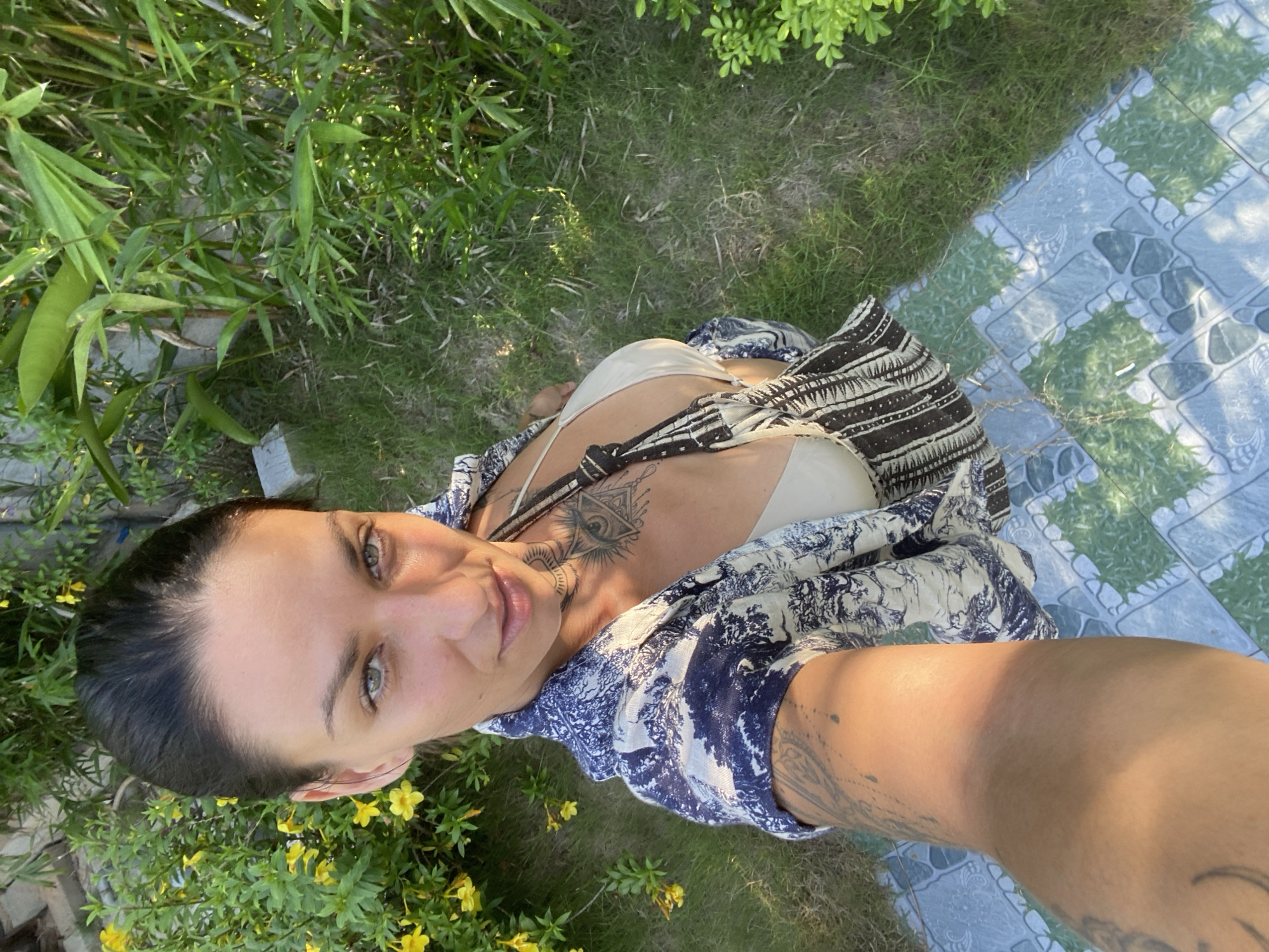 An overhead, full-body, selfie-style shot of a woman with dark hair pulled back stands outdoors in what appears to be a garden or yard in Mũi Né, Vietnam. She is looking at the camera, with her head tilted slightly. The woman is wearing a cream-colored bikini top and an open, short-sleeved shirt with a blue and white pattern. A dark-colored bag with a patterned strap hangs from her shoulder. She has numerous tattoos visible on her chest, arm, and leg. The background consists of green grass, various plants, and yellow flowers. A tiled walkway is visible in the lower portion of the image. The lighting suggests it is daytime, with sunlight casting shadows.
