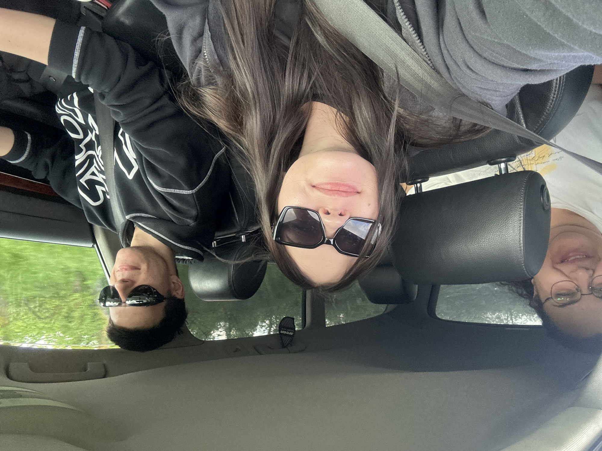Three people are inside a car. The person on the left wears a black hoodie with white graphics and sunglasses. The person in the middle has long brown hair and wears black rectangular sunglasses. The person on the right wears glasses and a white shirt. All three are wearing seatbelts. Greenery can be seen through the car windows. The interior of the car is primarily gray. The location is not Konstantinovo, Bulgaria.