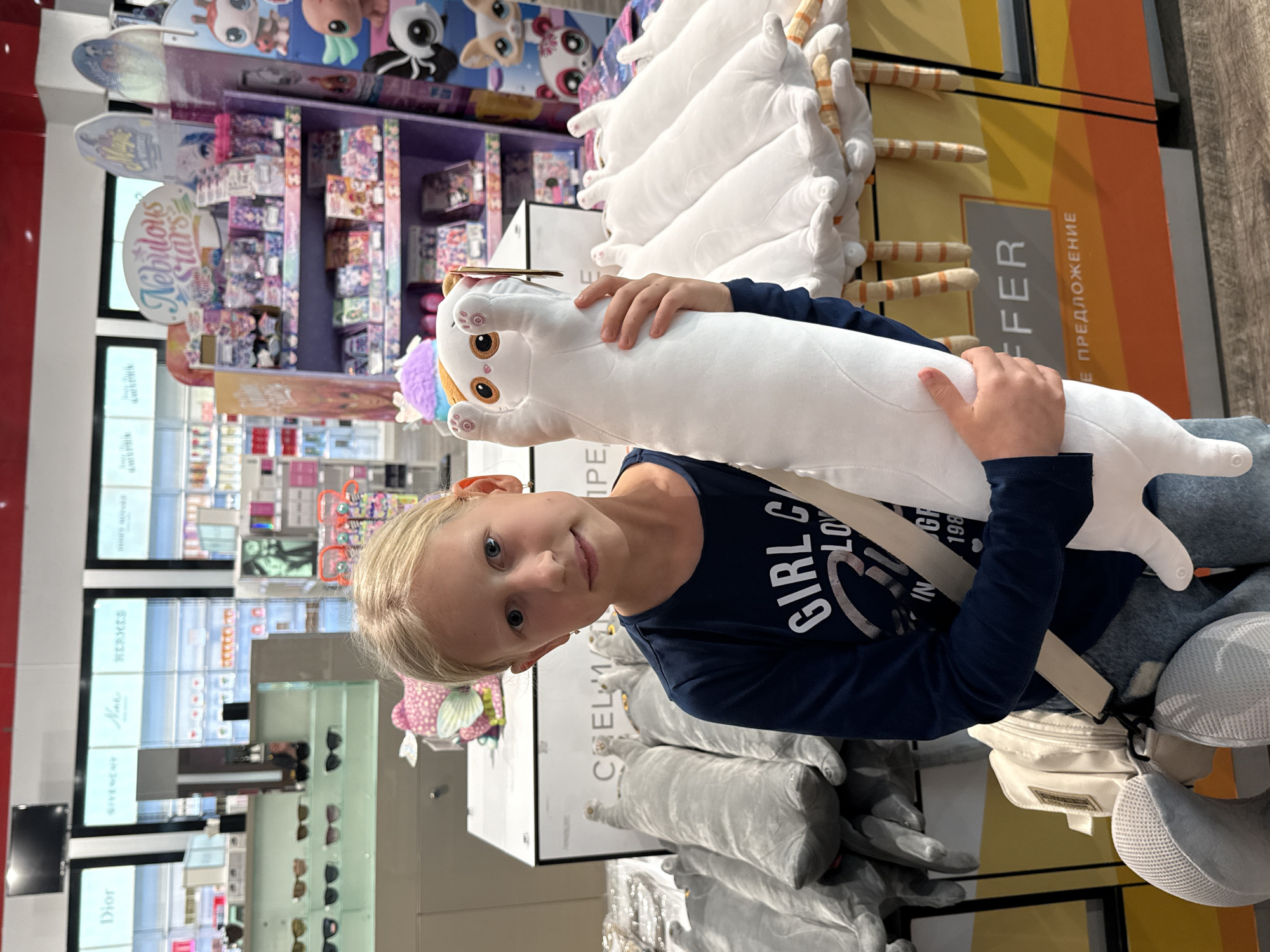 A young girl holds a white, plush, cat-shaped pillow in a store, presumably in Ufa, Russia, as the word "ПРЕДЛОЖЕНИЕ" in Russian is visible on a display in the background. The store appears to sell a variety of items, including toys and cosmetics, with brands like Dior and Givenchy displayed. The girl wears a dark blue, long-sleeved shirt, jeans, and a white bag slung over her shoulder. She is looking directly at the camera with a slight smile. Other plush toys are displayed around her, including additional white cat-shaped pillows and gray plush toys.